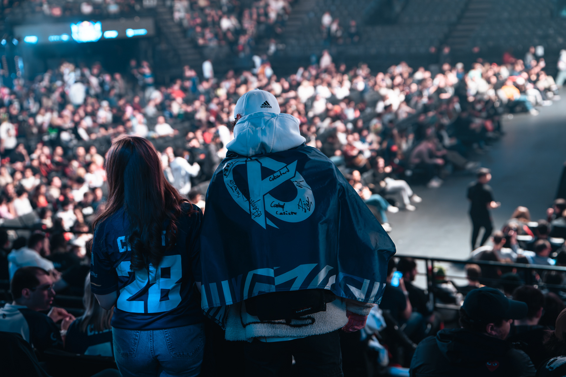 Vue de dos de deux fans à l'Accor Arena, l'un portant un drapeau de la Karmine Corp dédicacé sur les épaules face à une salle comble.