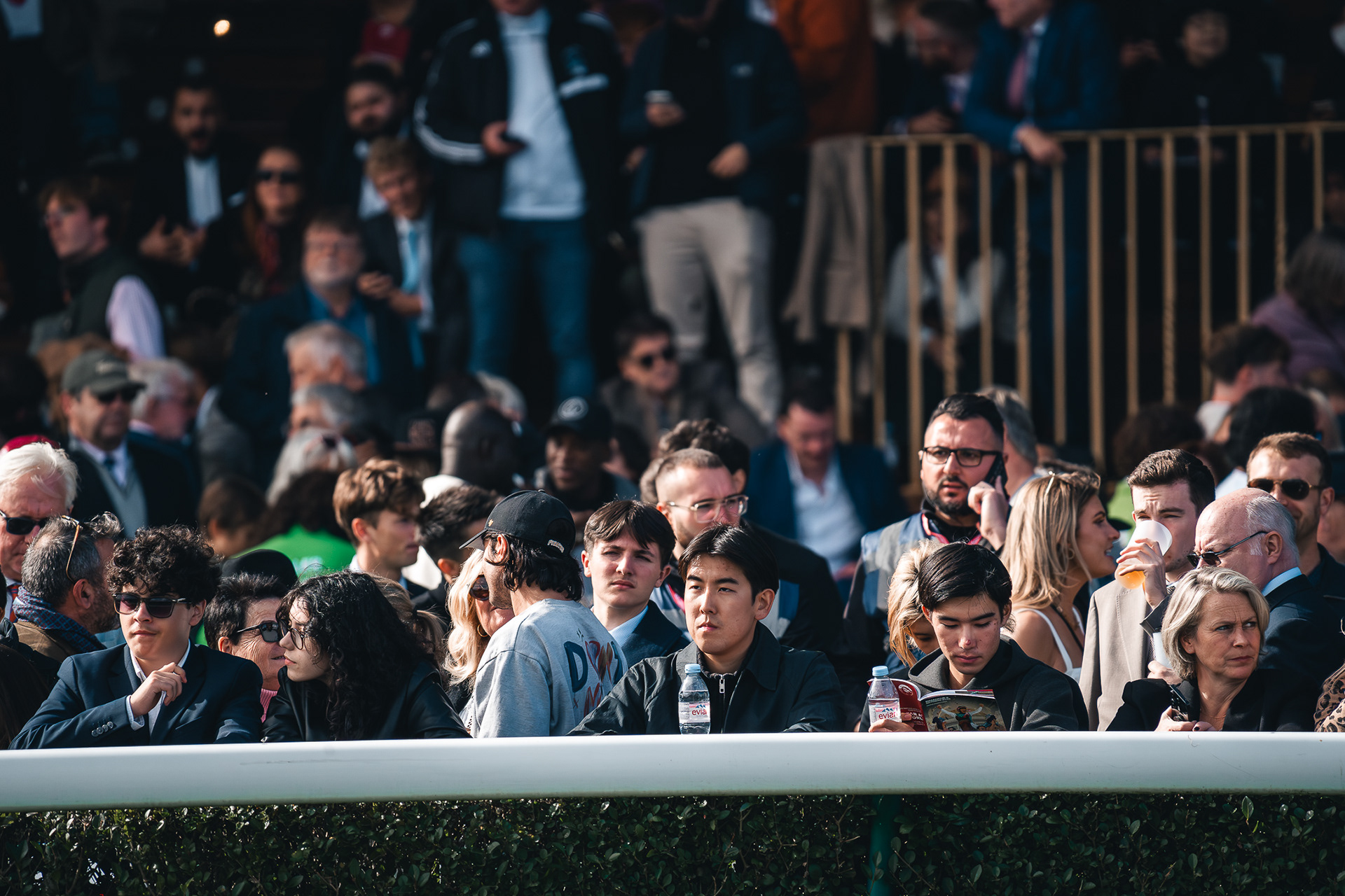 Gros plan de Sabri Kaced sur la foule de spectateurs attentifs derrière la barrière de la piste, certains lisant le programme des courses.
