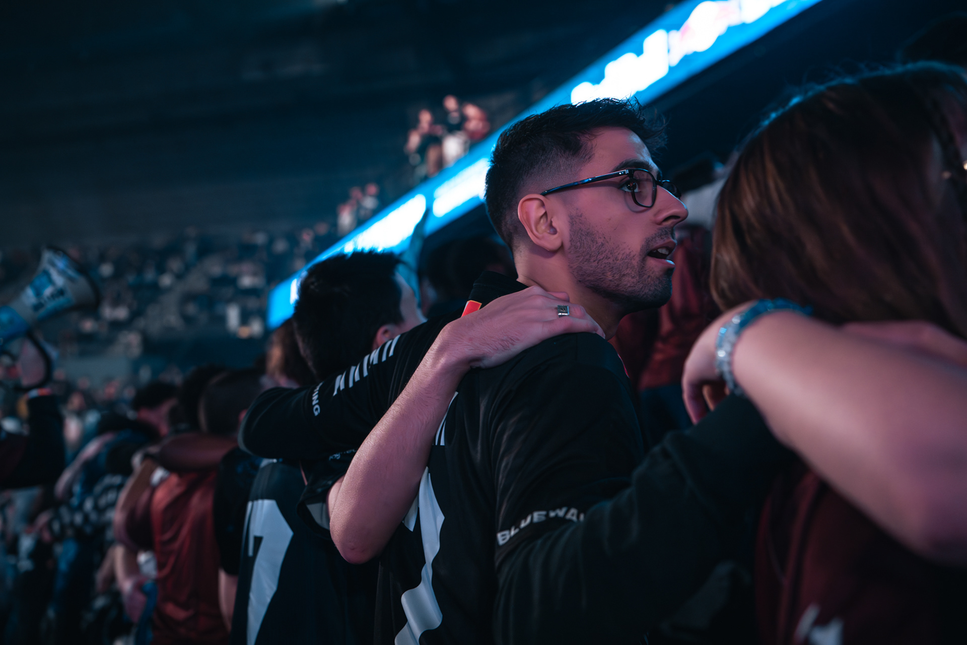 Portrait de profil d'un supporter avec lunettes dans une foule soudée, bras dessus bras dessous, lors d'une compétition de League of Legends.