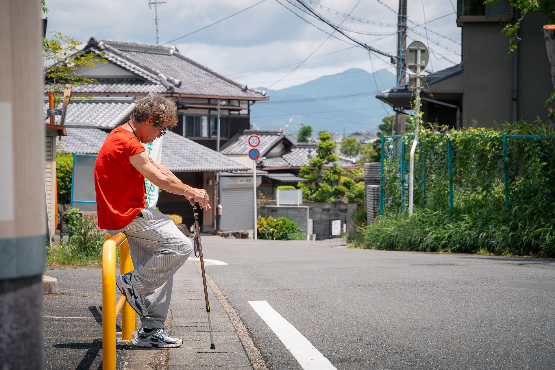 Portrait environnemental réalisé par Sabri Kaced d'un homme s'appuyant sur une barrière jaune dans une rue résidentielle avec des montagnes en arrière-plan.