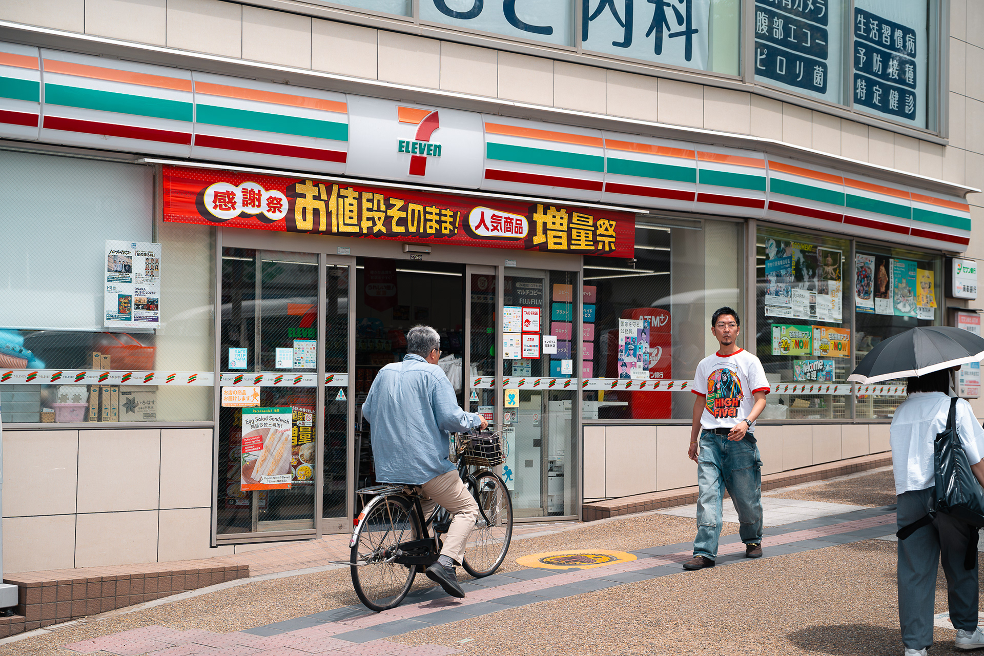 Scène de rue capturée par Sabri Kaced devant une supérette 7-Eleven au Japon avec un cycliste et un passant qui traverse.