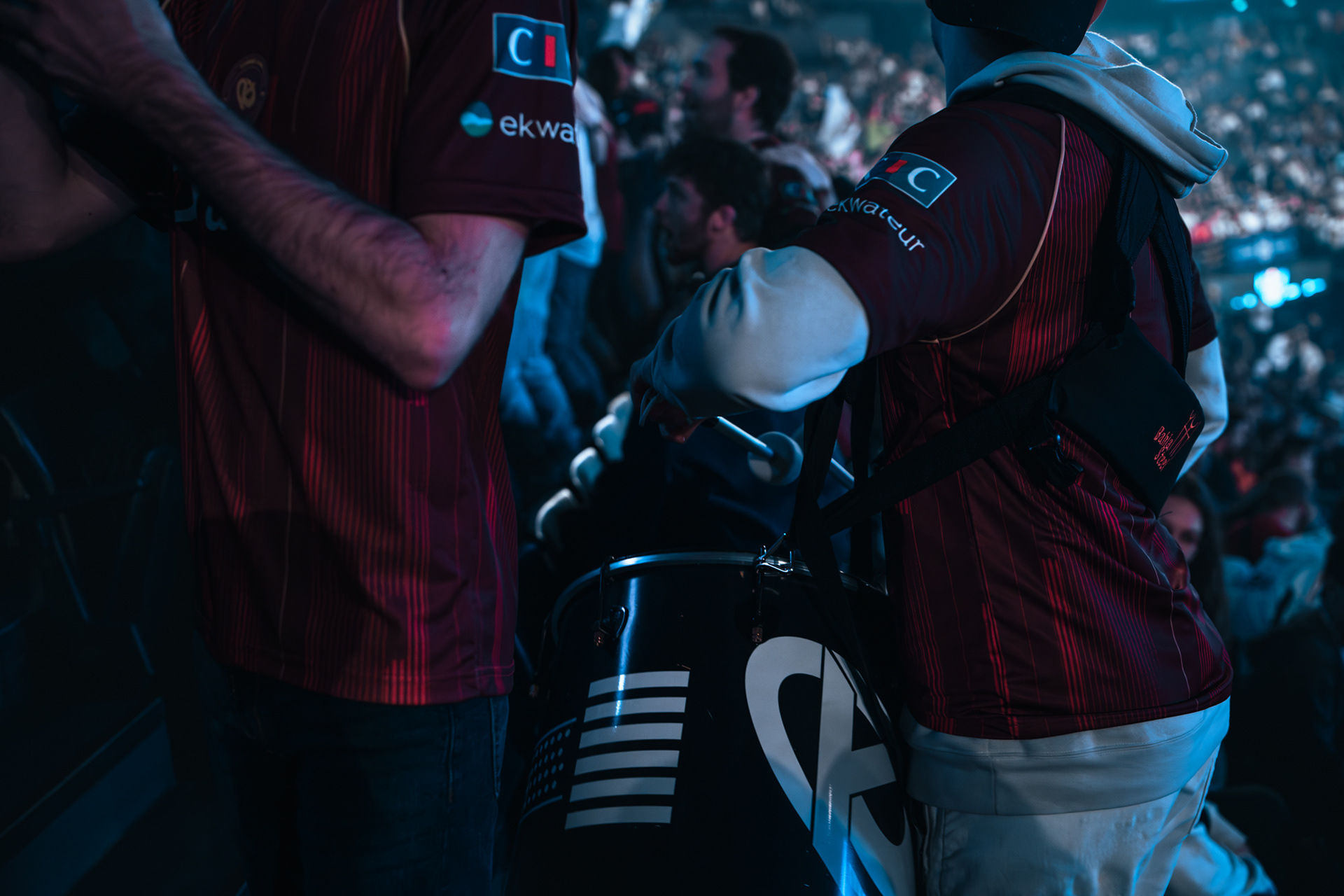 Gros plan sur un supporter de la Karmine Corp portant un maillot bordeaux et actionnant un tambour marqué du logo KC pour animer le stade.