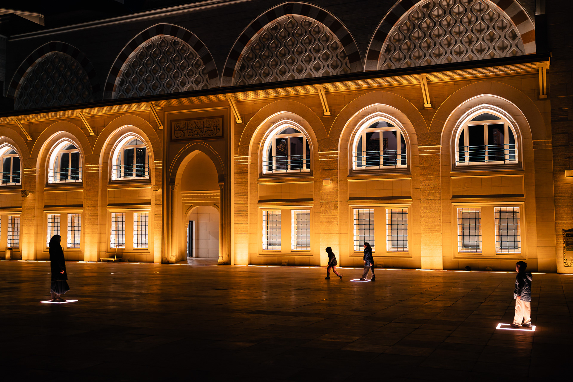 Vue nocturne de la façade illuminée de la mosquée Camlica à Istanbul, prise par Sabri Kaced, avec des silhouettes au premier plan devant les arches éclairées.
