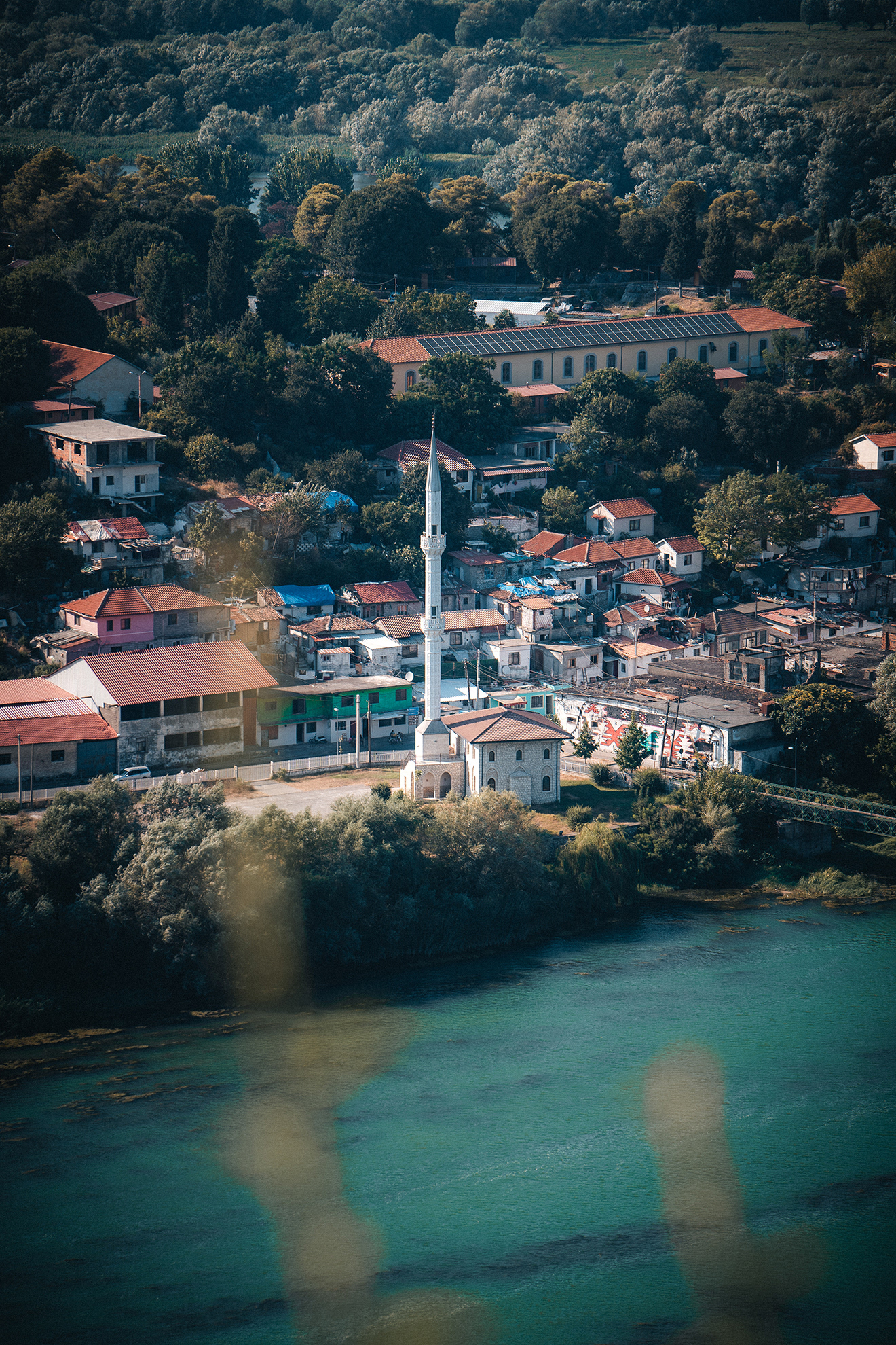 Vue panoramique en hauteur de la ville de Shkodër en Albanie, montrant une mosquée blanche, les toits rouges et la rivière Buna entourée de végétation. Photo : Sabri Kaced.