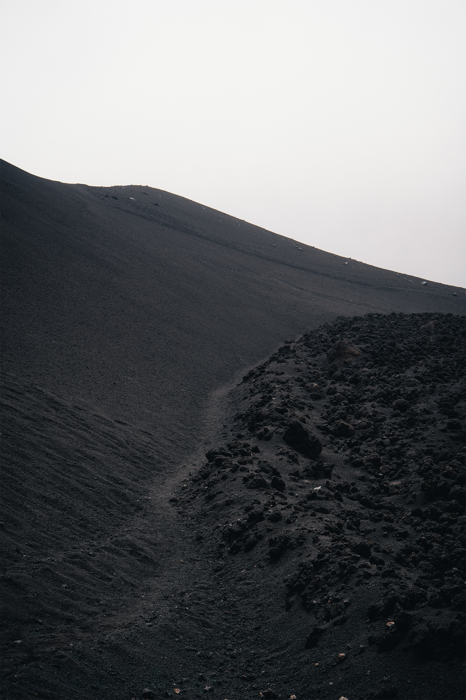 Sentier de randonnée sinueux tracé dans la cendre noire volcanique sur les pentes de l'Etna en Sicile, sous un ciel blanc minimaliste.