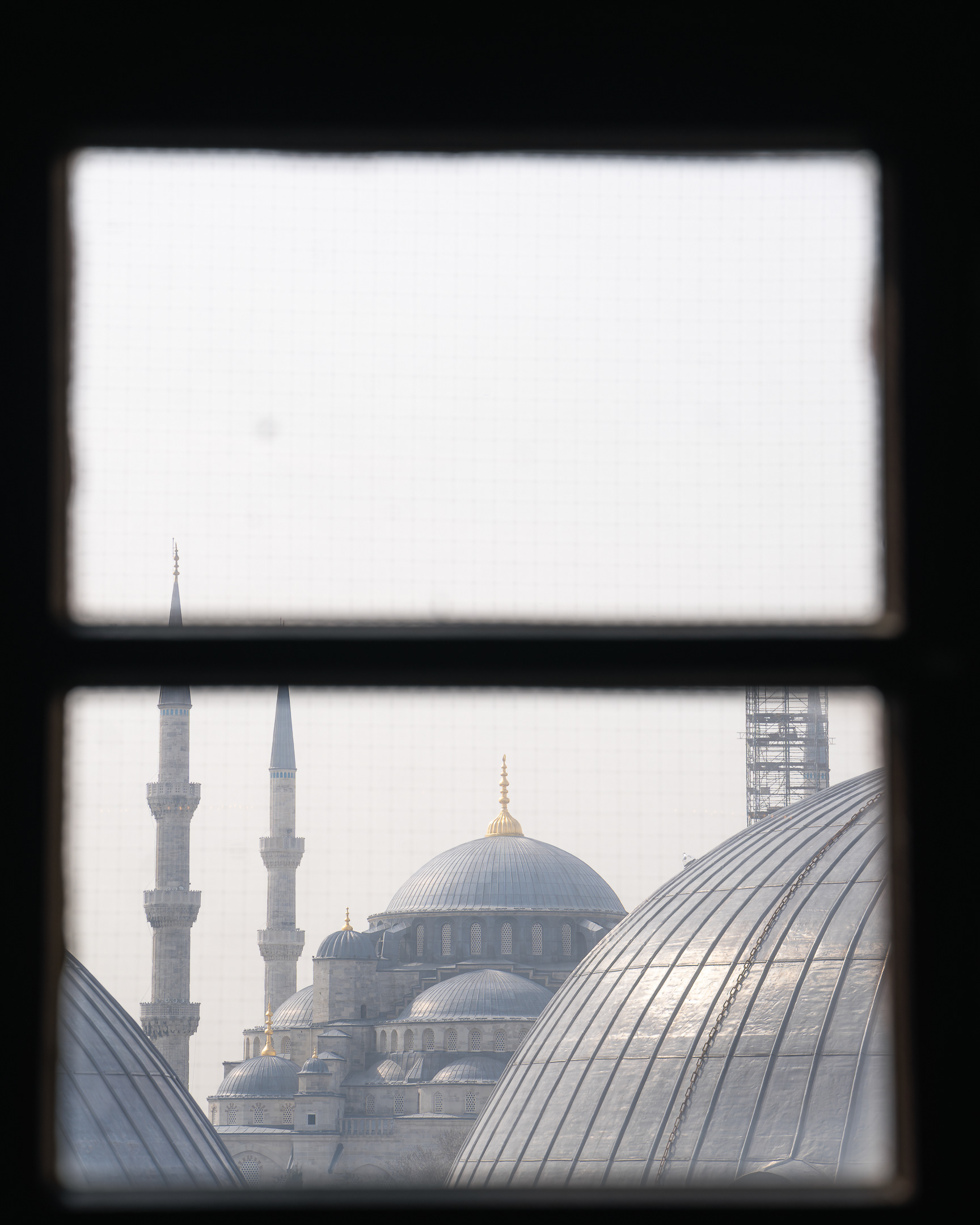 Composition de Sabri Kaced à travers un cadre de fenêtre montrant les dômes gris et les minarets de la mosquée Sultan Ahmed dans la brume d'Istanbul.