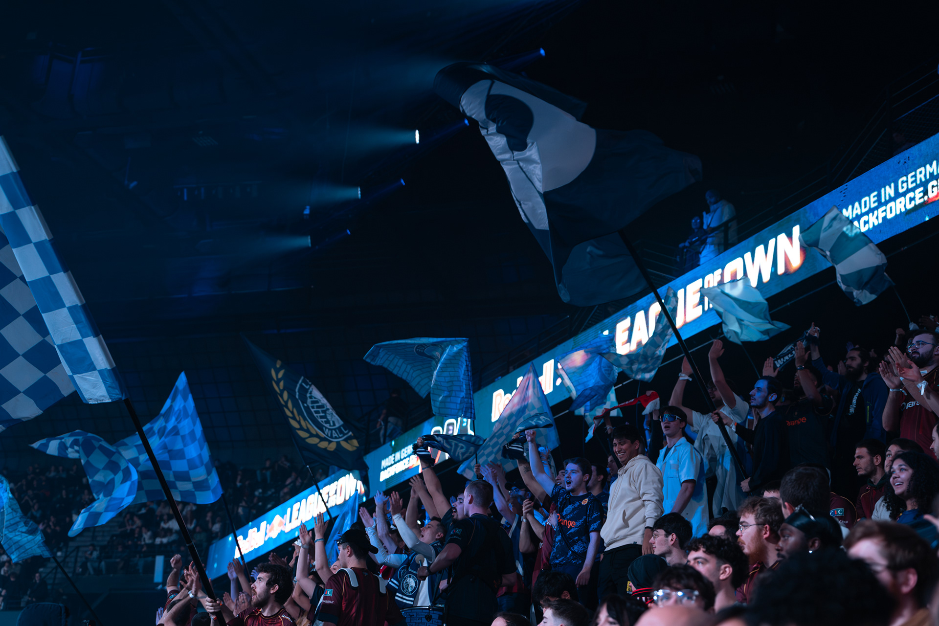Vue panoramique de l'arène esport avec des rangées de supporters levant les bras et de grands drapeaux bleus en mouvement.