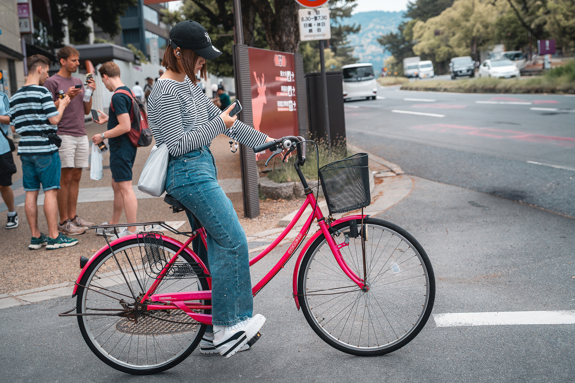 Photo de style lifestyle signée Sabri Kaced représentant une jeune femme de profil sur un vélo rose, consultant son téléphone dans une rue urbaine.