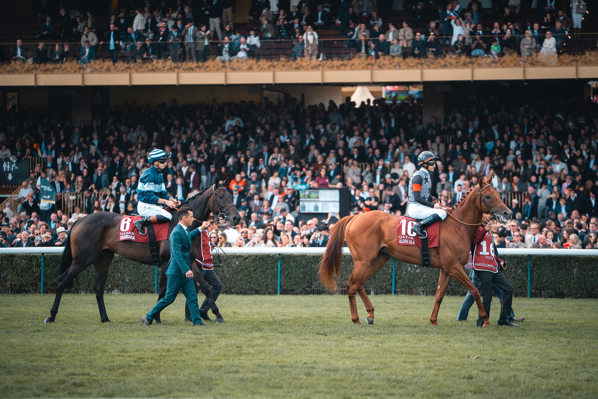 Photographie de Sabri Kaced : deux jockeys en tenue de course mènent leurs chevaux au pas devant des tribunes bondées lors d'un événement hippique.