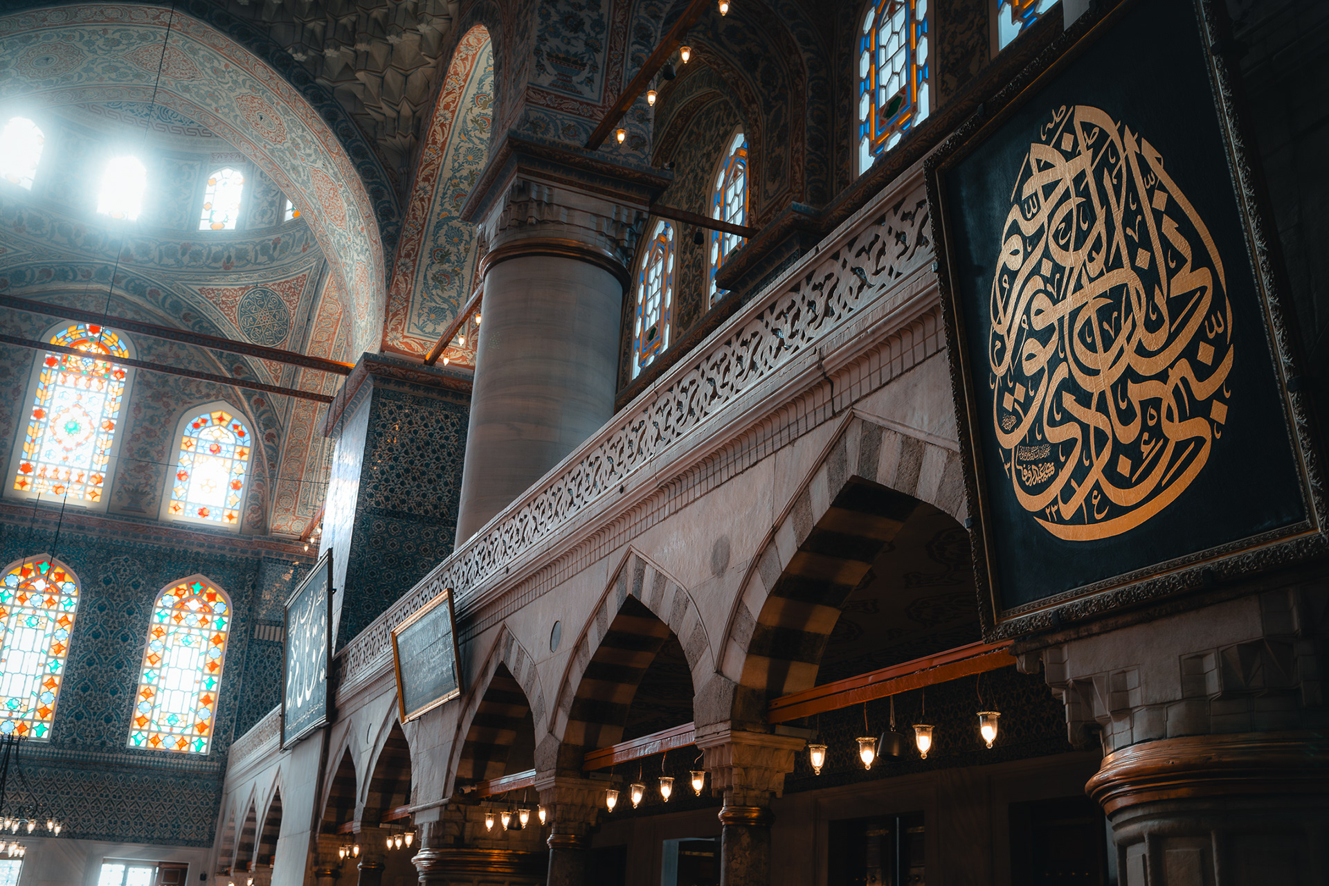Détail architectural capturé par Sabri Kaced présentant des arches en pierre, des panneaux de calligraphie dorée et des luminaires suspendus traditionnels.