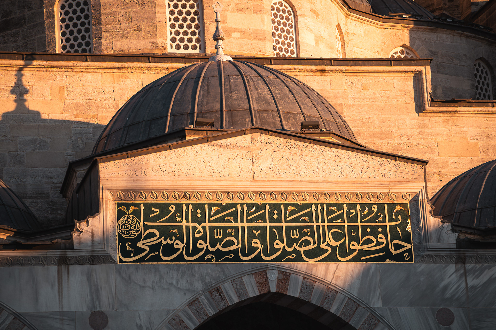 Gros plan sur une inscription calligraphique dorée au-dessus d'une porte de mosquée, illuminée par la lumière chaude du couchant, par Sabri Kaced.