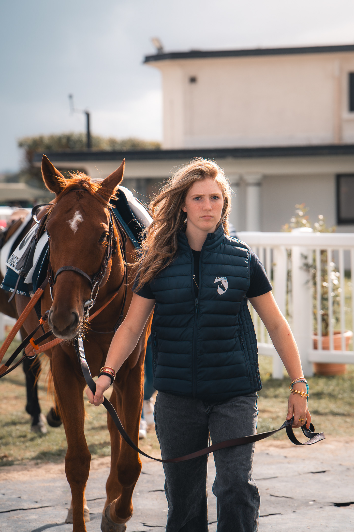Portrait par Sabri Kaced d'une jeune femme en veste sans manche tenant la bride d'un cheval alezan marqué d'une liste blanche sur le front.