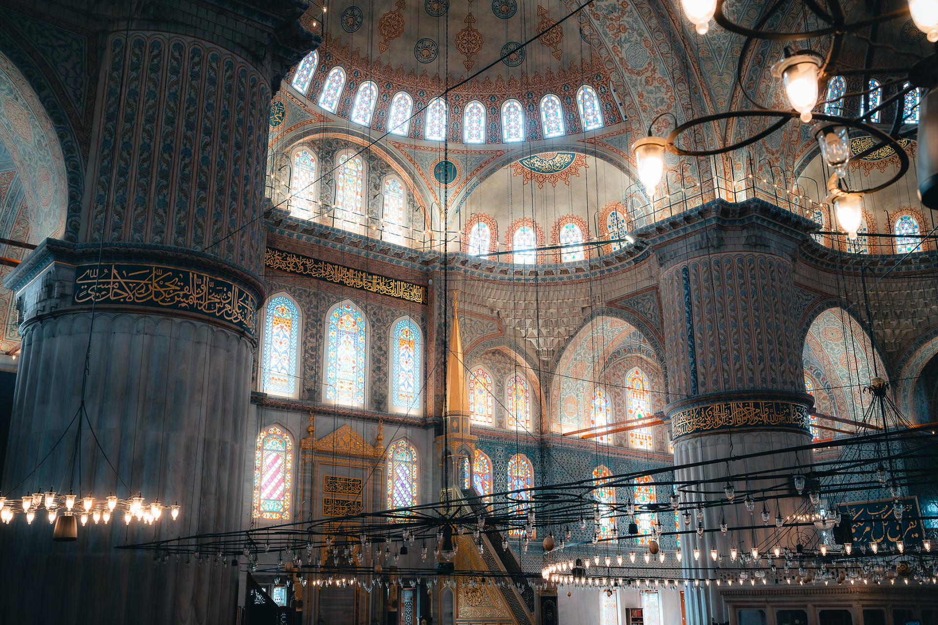 Photographie d'intérieur de la Mosquée Bleue par Sabri Kaced, mettant en avant les vitraux colorés et les piliers massifs décorés de calligraphies arabes.