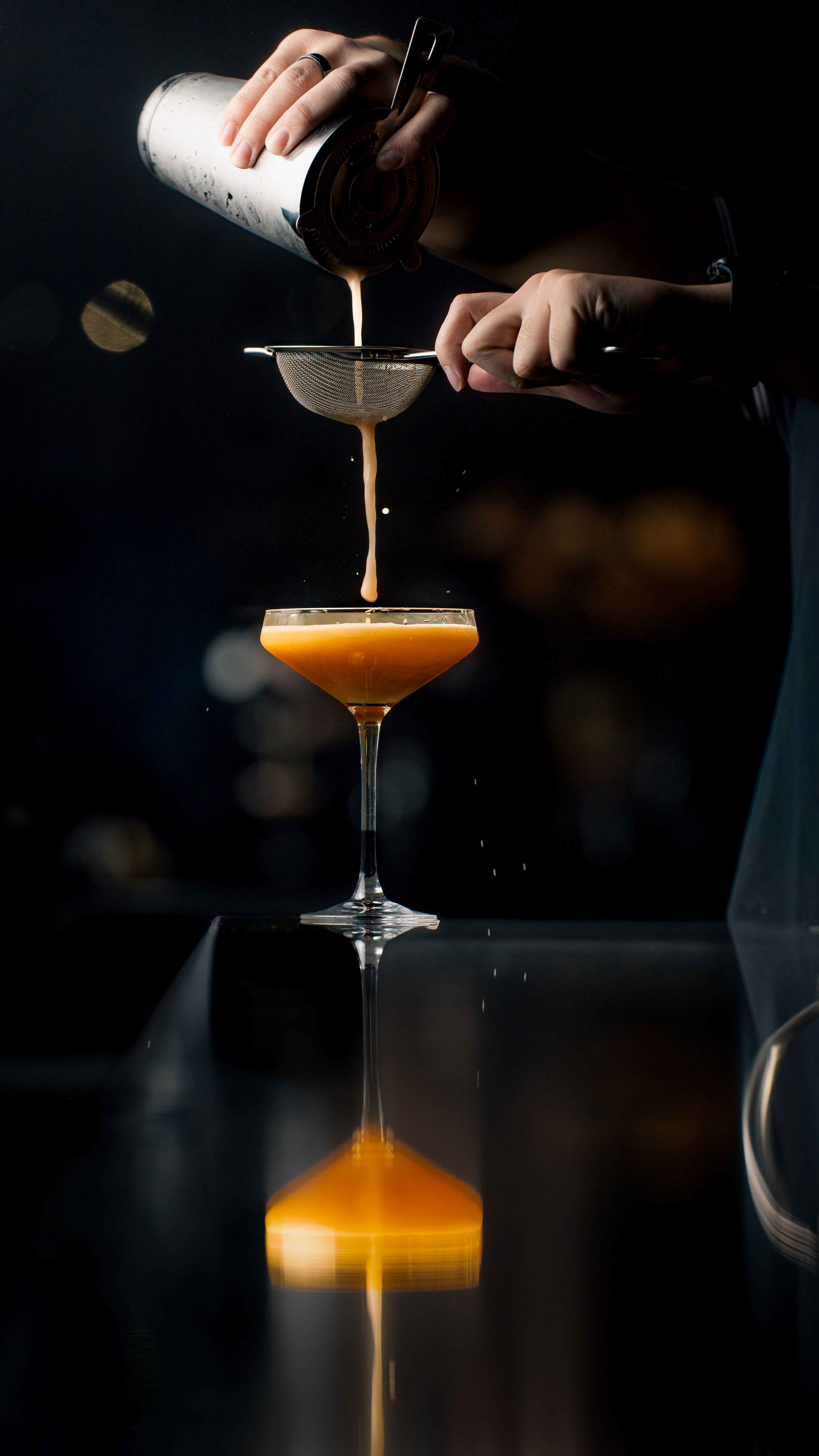 High-speed action shot of a bartender straining a vibrant orange cocktail into a coupe glass with a perfect reflection, luxury beverage photography for Toronto nightlife and spirits brands.