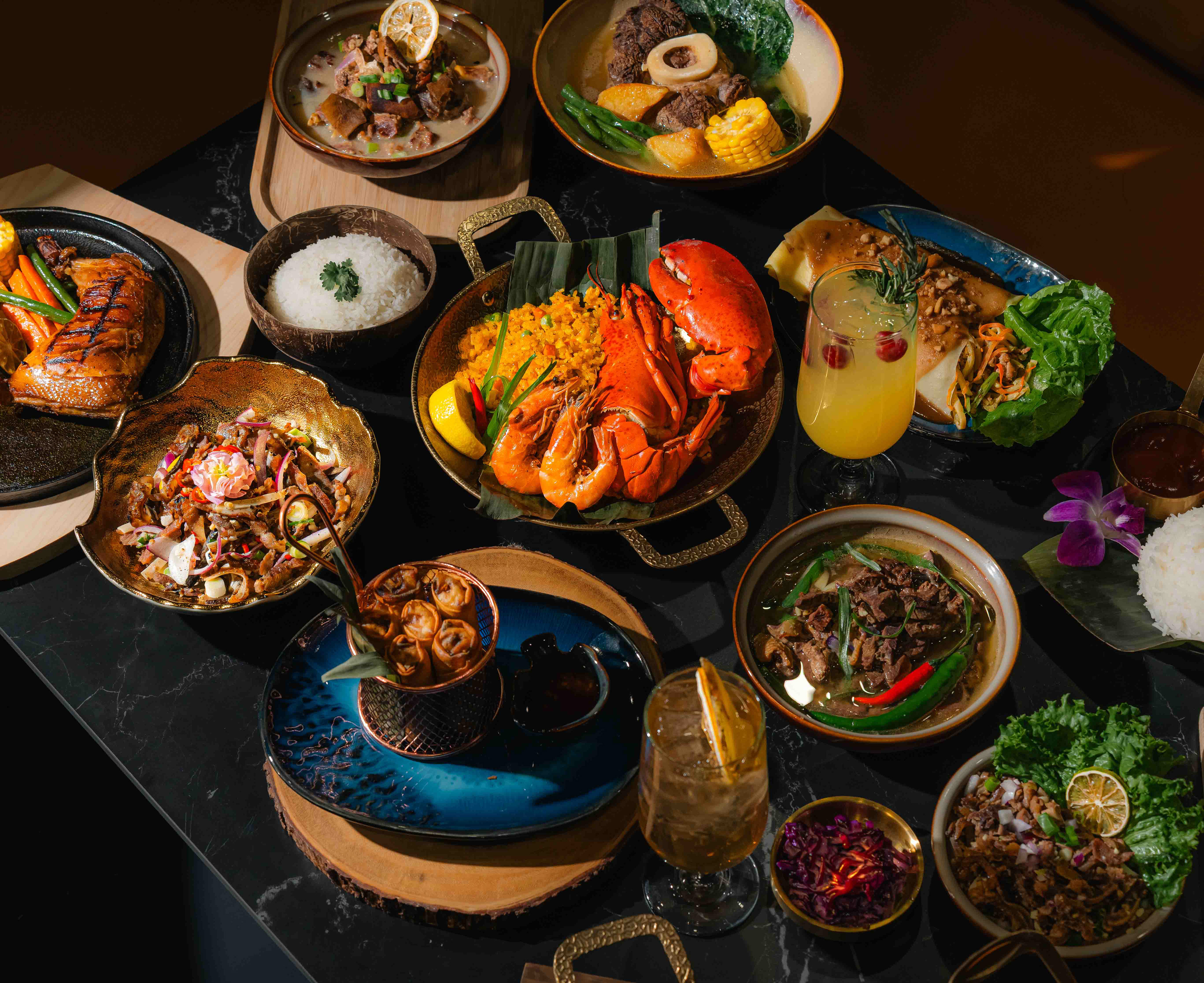 A luxury Filipino-inspired feast featuring lobster, paella, sisig, and tropical cocktails, photographed by Ash, a professional food and drink photographer in Toronto. This overhead group shot showcases high-end restaurant menu photography and commercial food styling in the GTA.