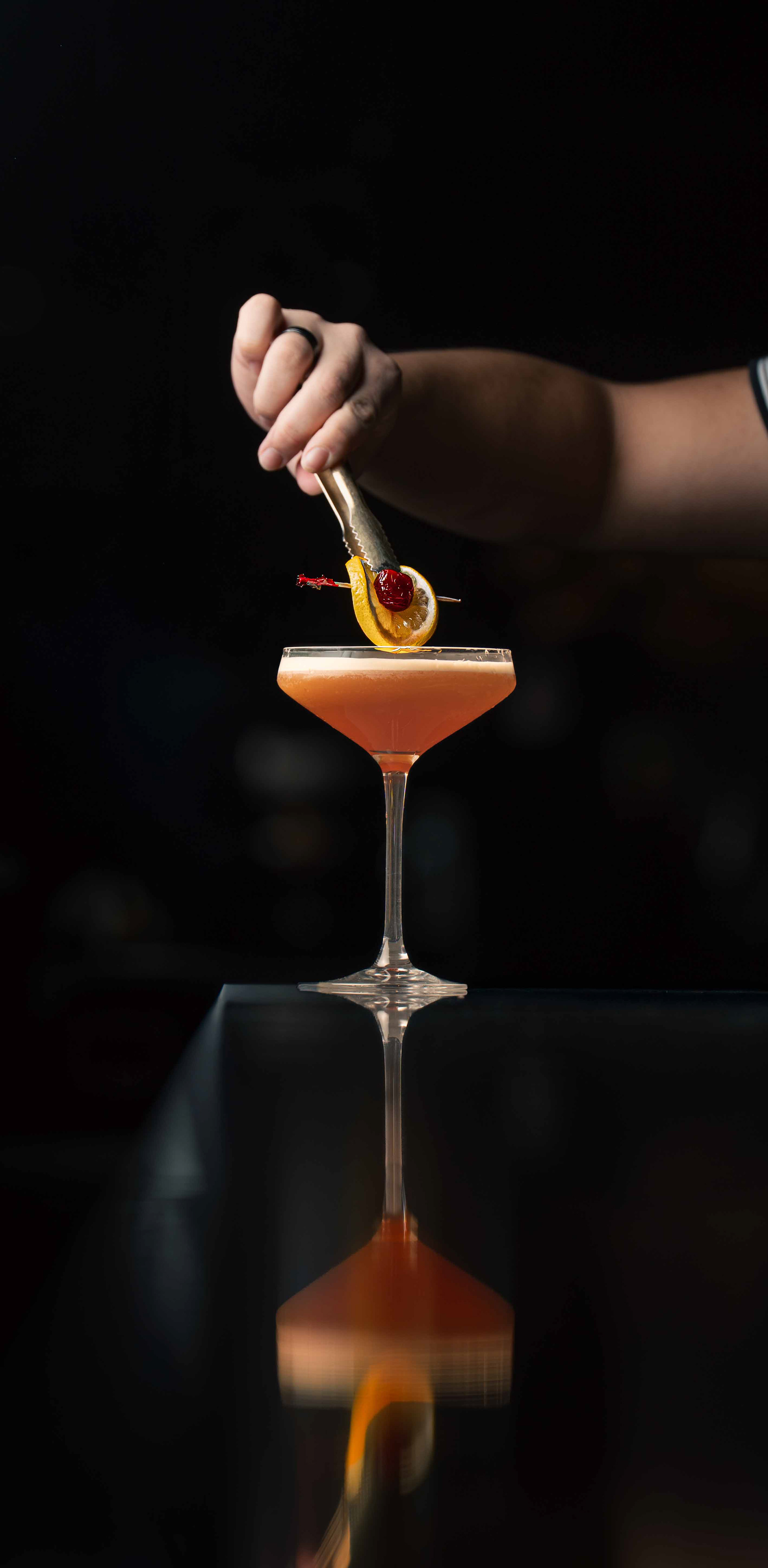 Close-up of a bartender using tongs to place a citrus and cherry garnish on a craft cocktail with a perfect reflection on a dark bar top. High-end beverage photography by Toronto videographer and food photographer Ash, showcasing precision lighting for cocktail bars and the hospitality industry.