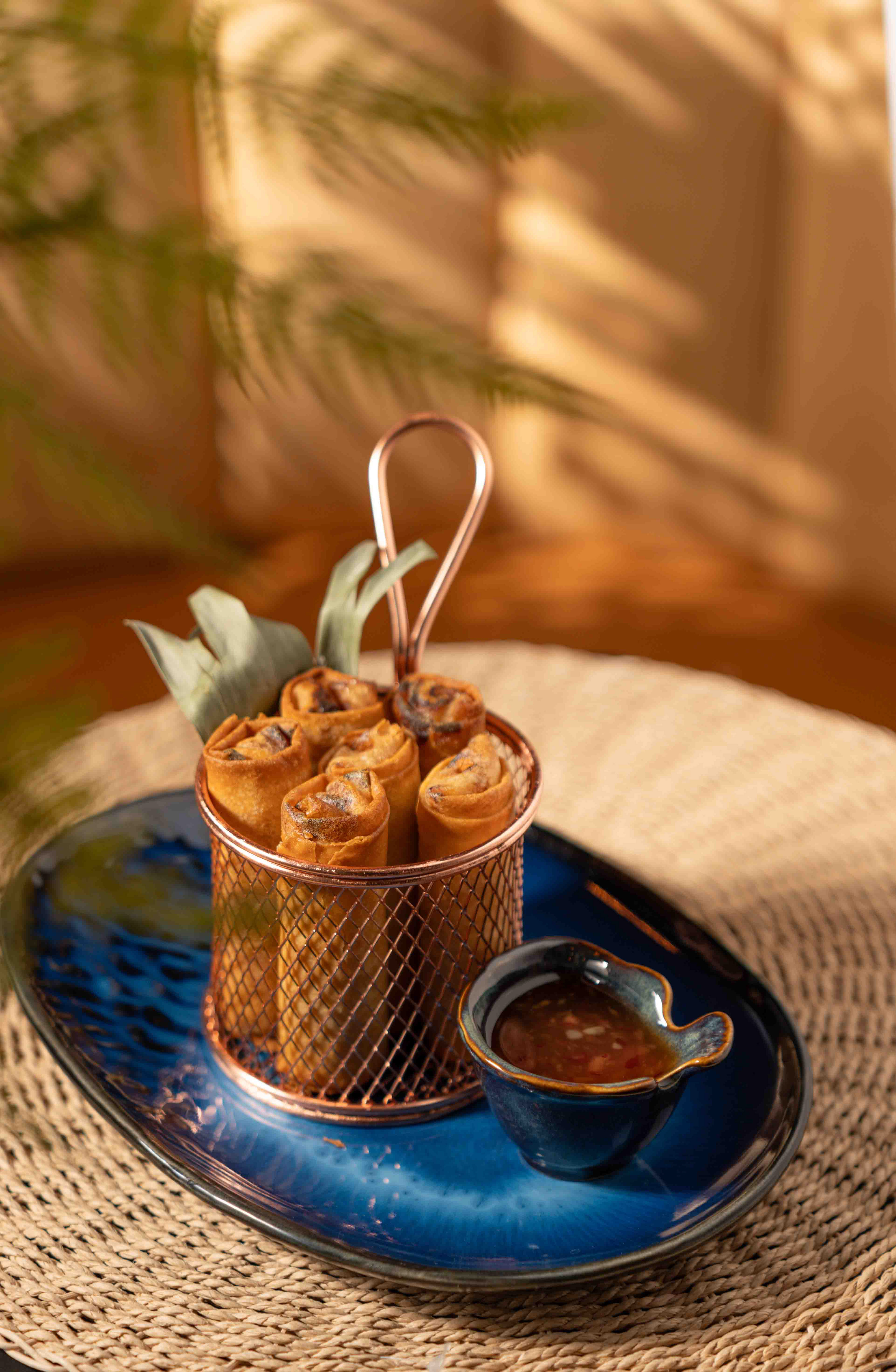 Gourmet spring rolls in a copper basket with dipping sauce, professionally captured by Ash, a food photographer and videographer in Toronto. The shot features warm, cinematic lighting with tropical leaf shadows, ideal for restaurant branding and commercial food marketing in the GTA.