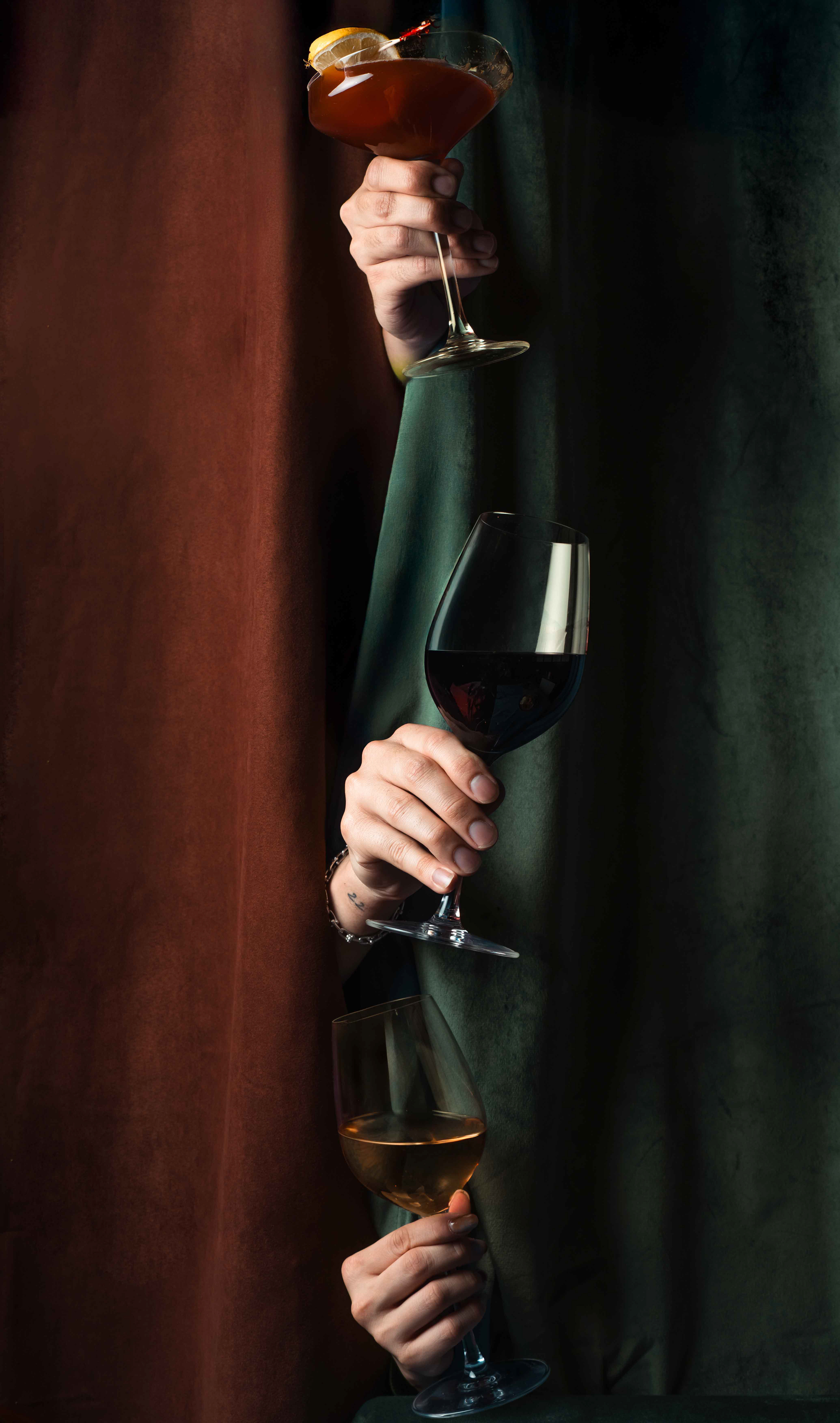 Artistic lifestyle beverage photography featuring multiple hands holding glasses of red and white wine against velvet curtains, professional hospitality imagery for Toronto bars and event venues.