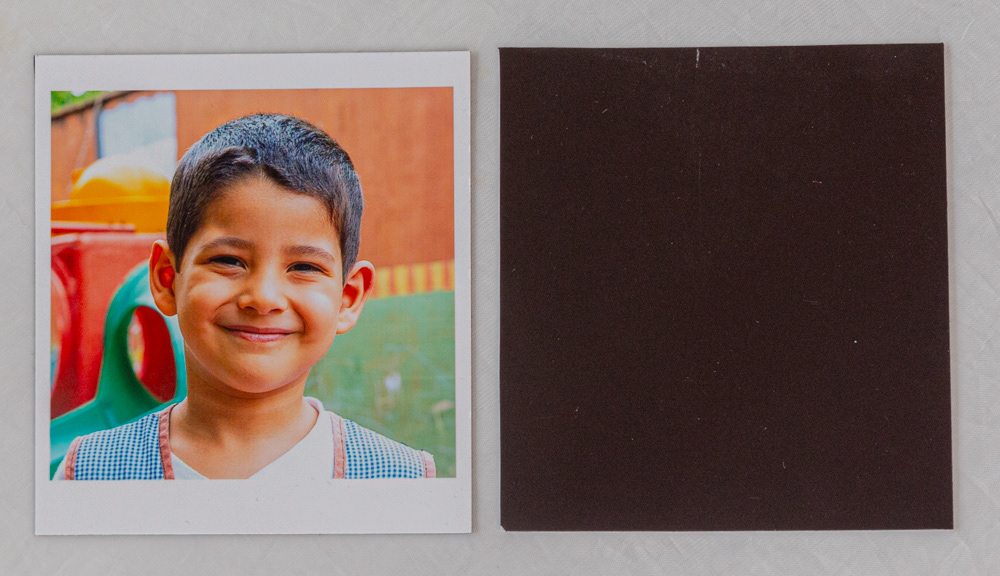 Foto estilo polaroid de 8x10 cm con imán en toda la superficie del dorso. Niño sonriendo en patio de jardín con juegos de fondo. Producto adicional 2026 de Retratos Escolares: recuerdo escolar magnético para heladera, fotografía con luz natural en CABA y Gran Buenos Aires.