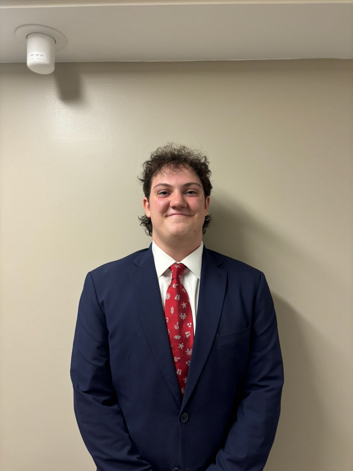 Image of Charlie Raibman standing against a tan wall. He is wearing a navy blue suit and a white dress shirt. He is wearing a red tie with nautical icons. He has curly brown hair.