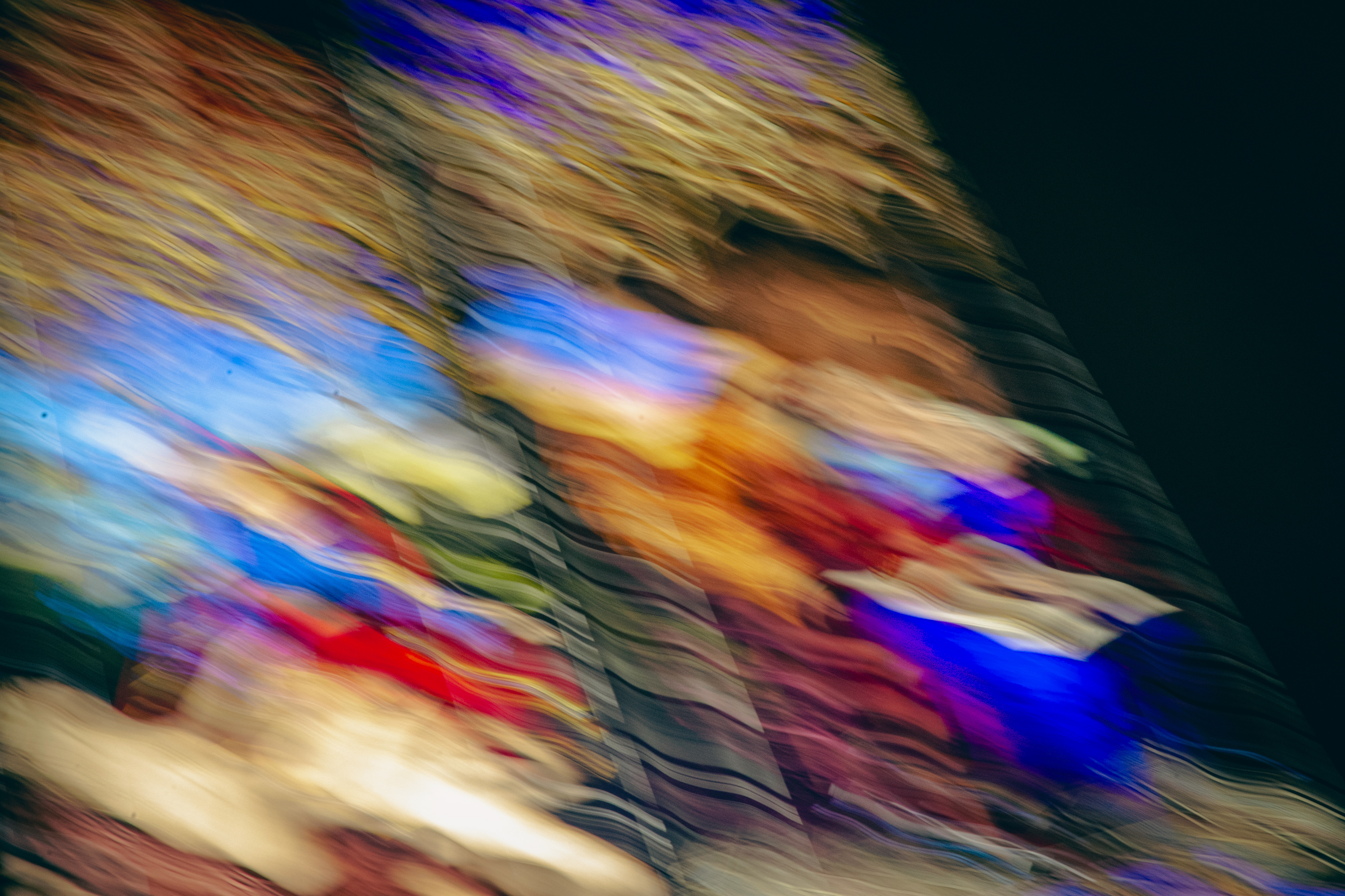 'Shimmering Liquid Light' (2025) A sweeping wash of vivid colour and shifting textures fills the image. Electric blues and deep purples streak diagonally across the scene, mingling with splashes of fiery red, sunlit yellows, warm oranges, and soft creamy whites. The colours appear to be smeared and stretched, forming fluid, wavy lines that ripple through one another, like reflections on rippling water. Bright, glowing areas of colour are interspersed with darker shaded bands, creating a richly layered effect. The texture suggests smooth glass or polished surfaces, and the interplay of light caught in these swirling patterns evokes the energy and intricacy of a stained glass window seen through a moving or watery lens. The overall impression is vibrant, dynamic, and immersive, surrounding the viewer with a kaleidoscope of colour and movement.