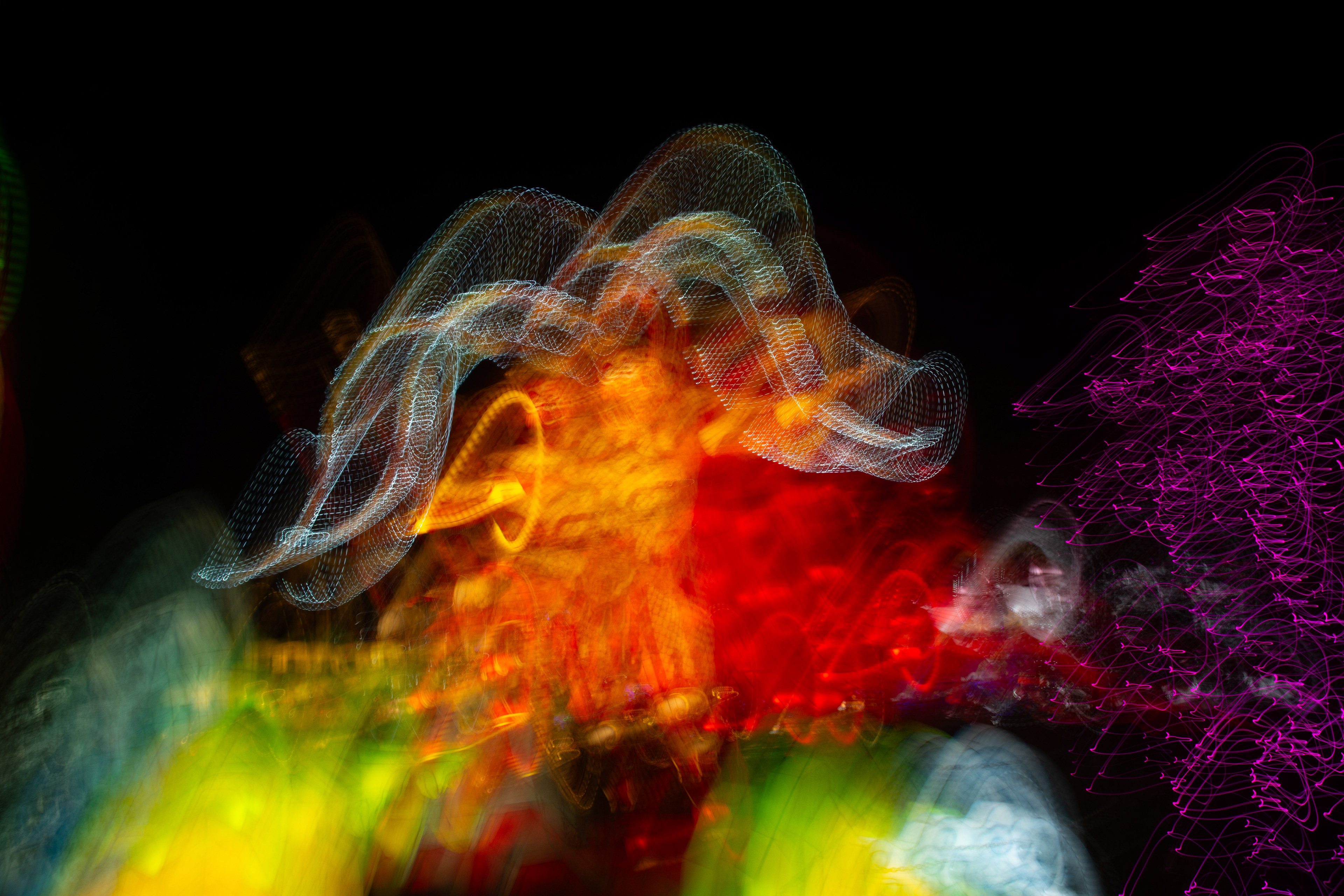 'Angel Wings' (2018). A celestial being emerges from the darkness, its ethereal form composed of swirling, luminous colours. Fiery orange and golden hues radiate from its core, surrounded by wisps of green and purple that seem to shimmer and pulse with life. The creature's form is both majestic and mysterious, as if it is weaving through the cosmos, leaving trails of light in its wake. Its presence is otherworldly, a mesmerising blend of colour and motion that captivates the imagination.