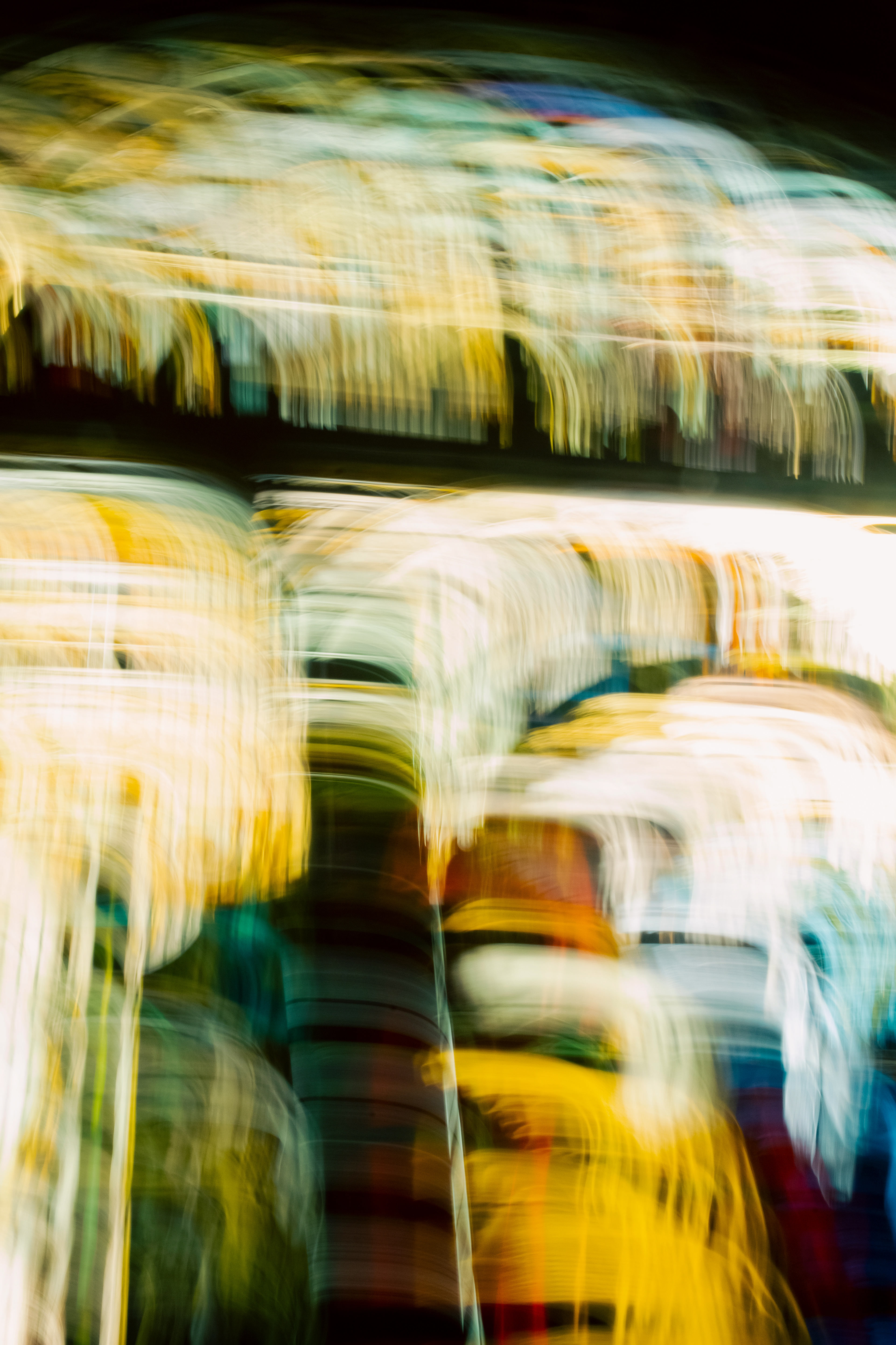 'Flowing Neon Arches' (2025) Streaks and swirls of colour dominate the image, creating a layered, visually textured effect. Vertical and slightly curved lines in yellow, white, teal, and a touch of rust red move across the frame, resembling painted brushstrokes or light trails. The yellow and white areas are brightest, almost glowing, while deeper greens and blues provide contrasting dark bands. The overall texture appears glossy and fluid—as if looking through glass smeared with wet paint or peering at city lights through a rain-soaked window. The blending of colours creates soft edges between streaks, resulting in a sense of depth and movement. The pattern is dense in some areas, especially at the top, giving a woven or tangled appearance, while in other places the space between lines allows light to shine through, adding to the rhythmic flow and layered complexity of the scene.