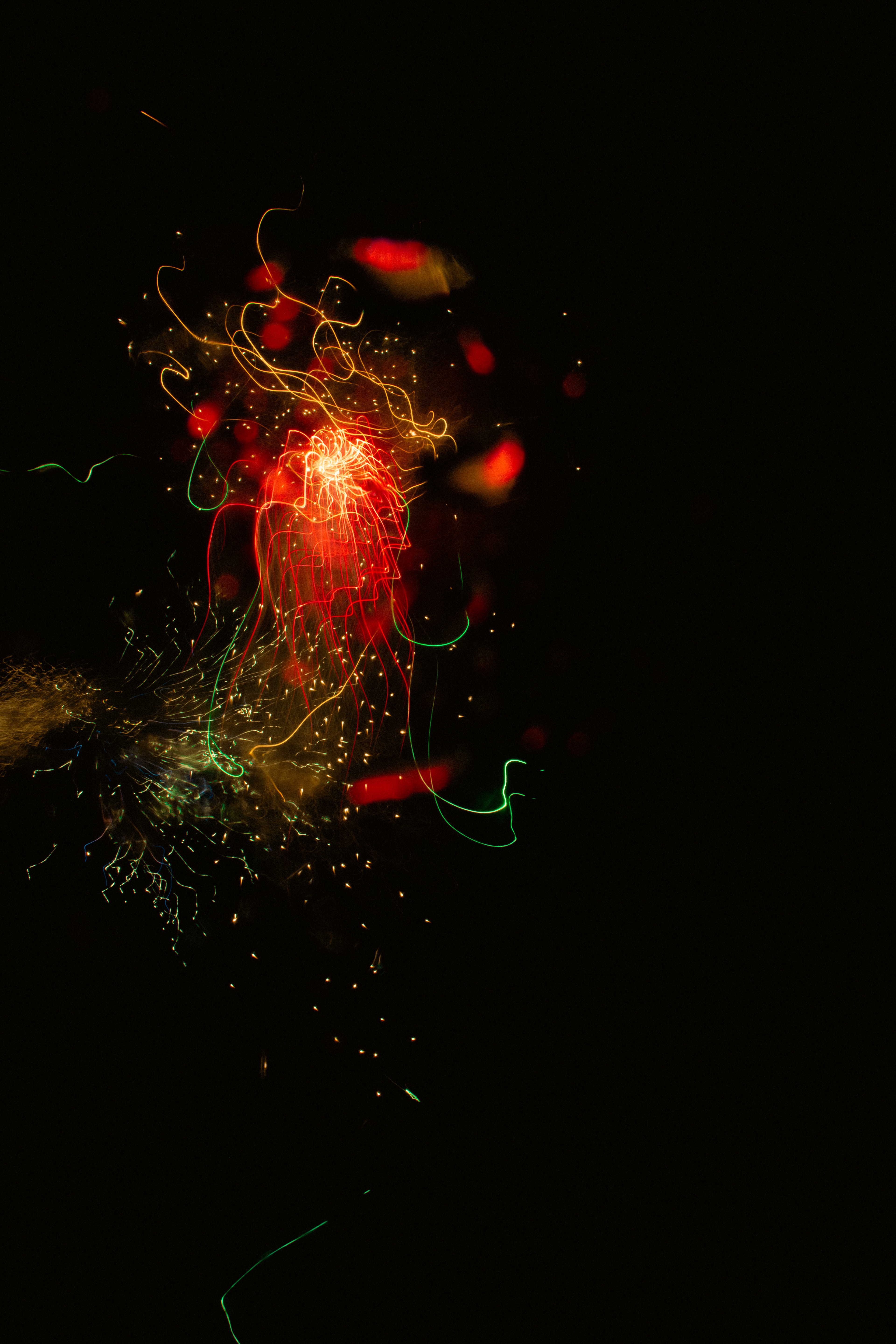 Sparks + Confetti I (2025) A burst of colorful light trails against a black background. Red, yellow, and orange lines swirl and intertwine at the center, with bright sparks radiating outward. Green and blue streaks weave through the chaos, creating a dynamic, fluid pattern. The glowing orbs and zigzag lines give the image a sense of movement and texture, resembling fireworks captured in mid-explosion.