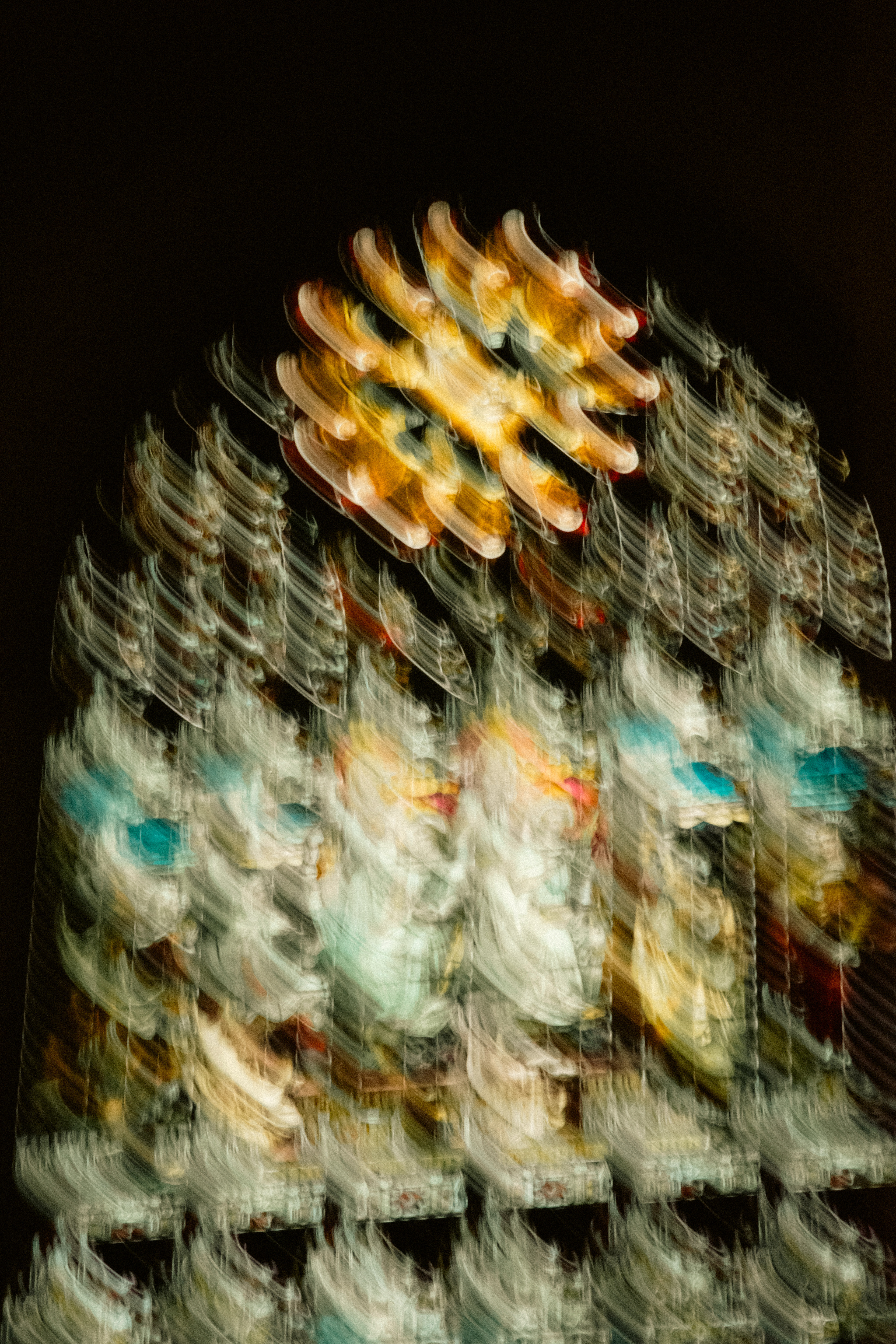 'Tactile Radiance' (2025). Wavy, blurred streaks of vibrant colour—yellows, oranges, blues, reds, and whites—form an abstract pattern. The glass texture appears soft and flowing, with no sharp edges or details, suggesting motion. Hints of stained glass structure and possible figures can be seen, but all outlines are melted together in sweeping, painterly strokes. The background is very dark, making the swirling colours pop. The top centre has three small, sharp white dots that stand out from the rest.