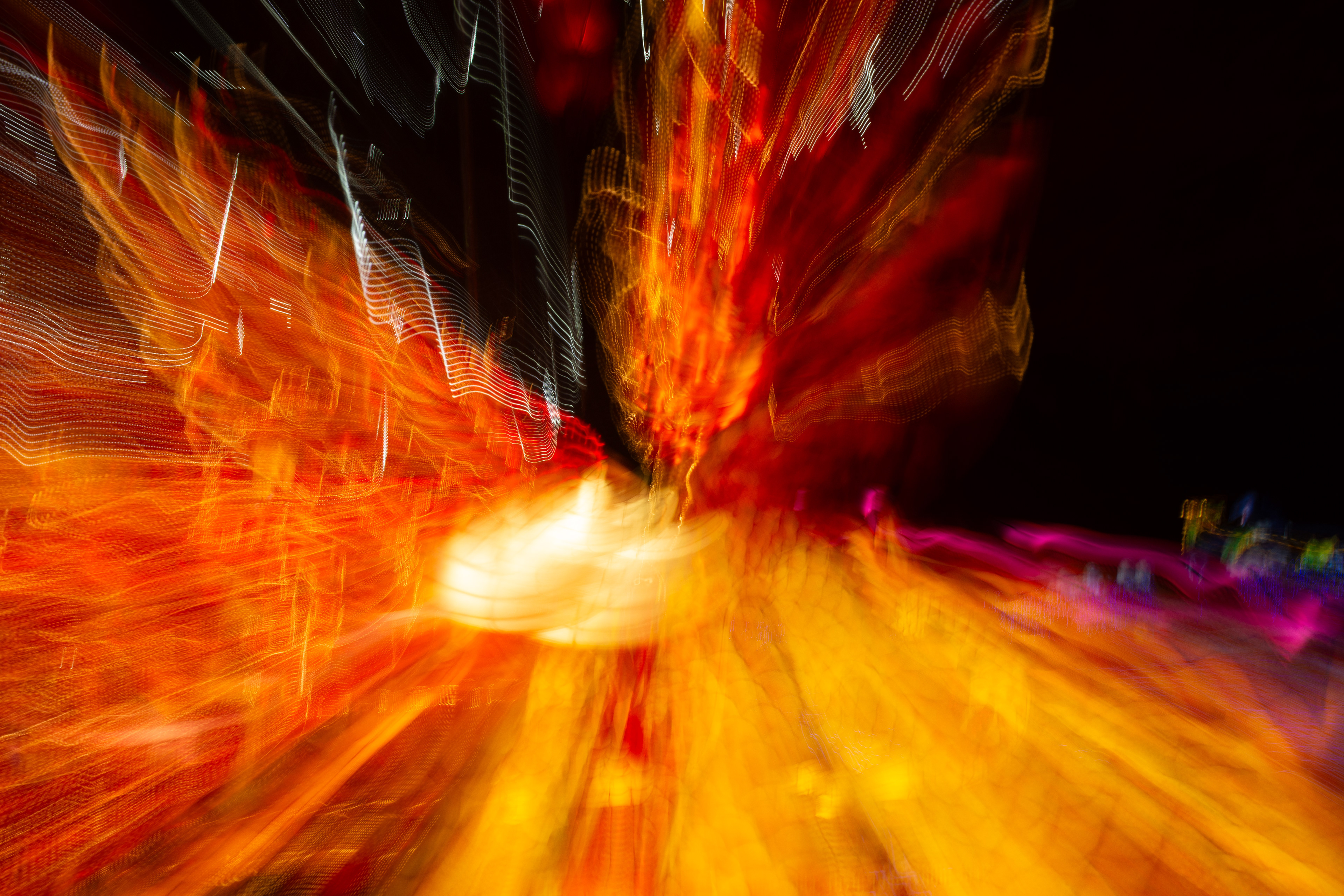 'Golden Phoenix' (2018). A chaotic, energetic explosion of color fills the frame, dominated by molten oranges, deep reds, and glowing yellows that ripple and twist like liquid fire. The light trails stretch and curl in every direction, forming tangled strands and wavering ribbons, as if the colors are being pulled outward in a sudden burst. Near the center, a bright, warm white glow flares like a small sun, diffusing into the surrounding heat-like hues. Subtle streaks of cool colors—thin purples, faint blues, and occasional silver-white lines—thread through the otherwise fiery palette, adding flashes of contrast. The overall texture feels fluid and swirling, like a long-exposure photograph capturing the movement of flames, sparks, and neon lights all blending into a single, vibrant storm of motion.