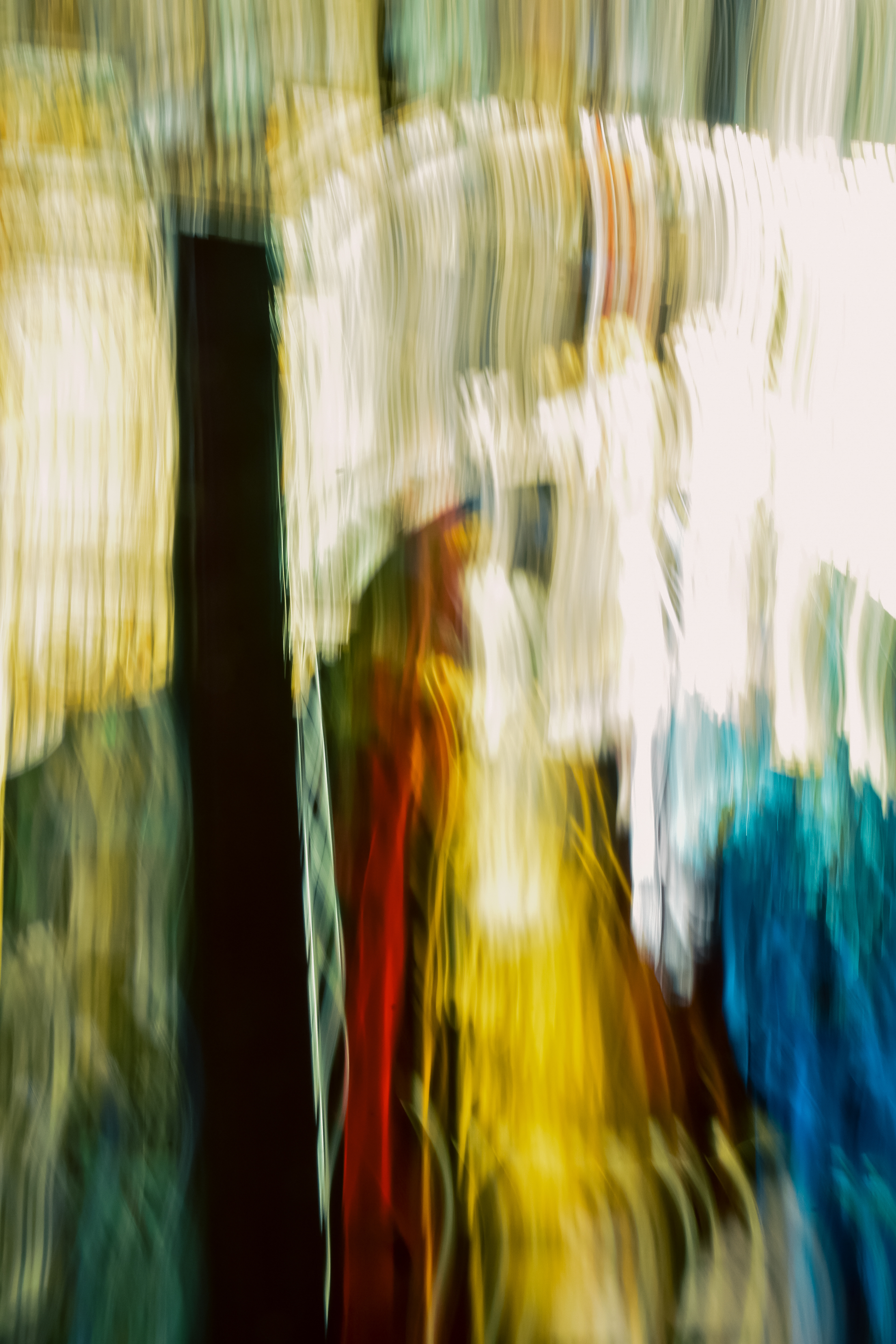 'Electric Cascades' (2025) Wavy streaks and bands of colour dominate the image, creating a strong sense of movement, as if the entire scene was captured while shaking or panning the camera. The textures are mostly smooth but appear layered and rippling, as if paint had been dragged or light reflected off water. The top area is lighter, filled with bright whites and golden yellows, almost glowing, with vertical lines appearing slightly curved and intersecting.    In the central section, a large dark vertical stripe contrasts with the brighter, adjacent hues. Around this, yellows blend softly with rich reds and a muted green, all forming elongated, twisting trails that fade into each other.    Toward the lower right, vivid blue and teal streaks create a deeper, cooler texture, the shades merging and separating, forming wispy, feathery patterns. Overall, the image feels fluid and dynamic, with the textures suggesting a mixture of silk, flowing water, and blurred neon lights—smooth in some parts, more jagged and erratic in others. The lower right corner contains a glossy, iridescent logo, which stands out from the rest of the textured, painterly surface.