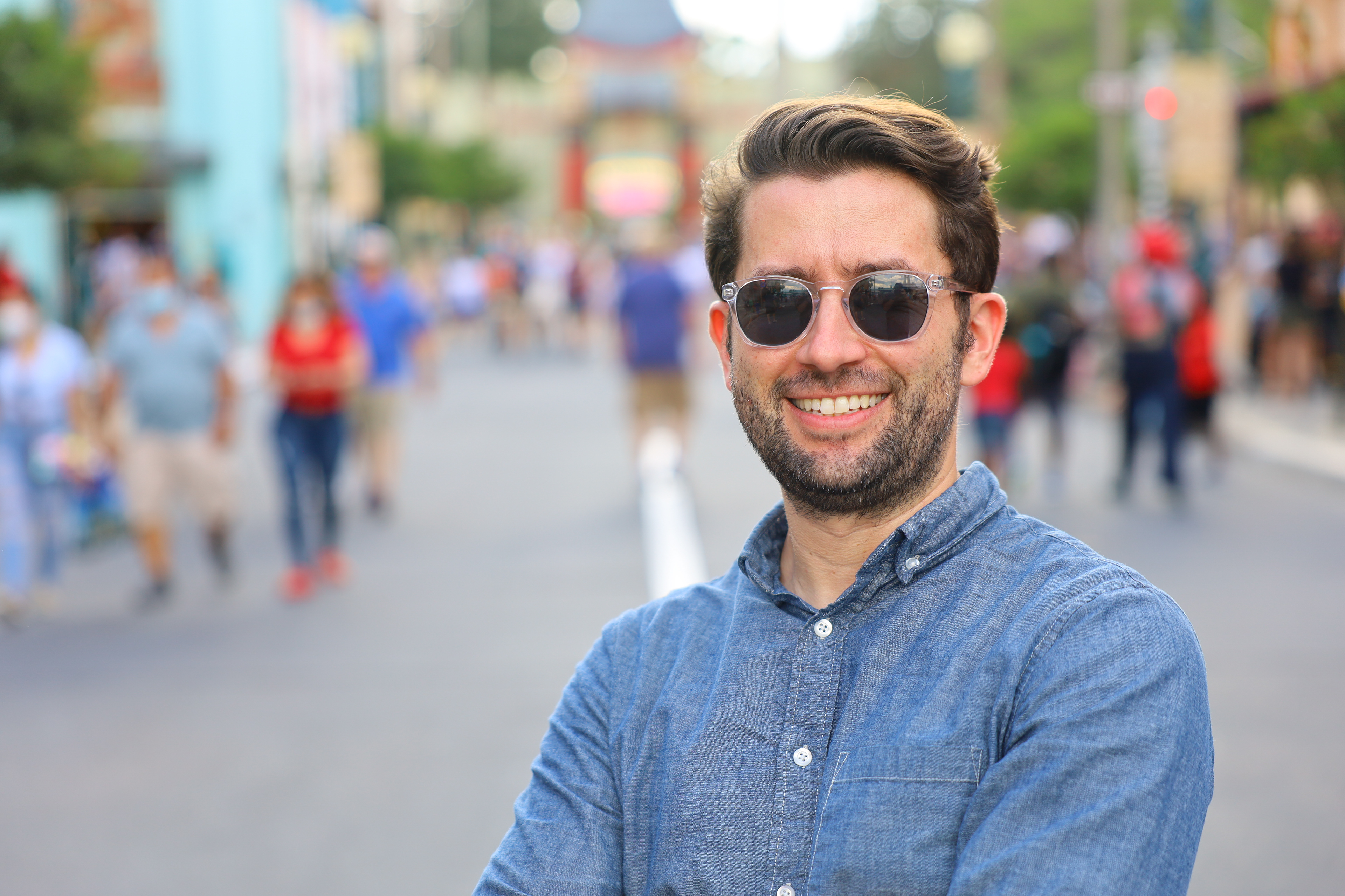 Man in a blue button up shirt and sunglasses smiles at the camera. People are in the background but very blury.