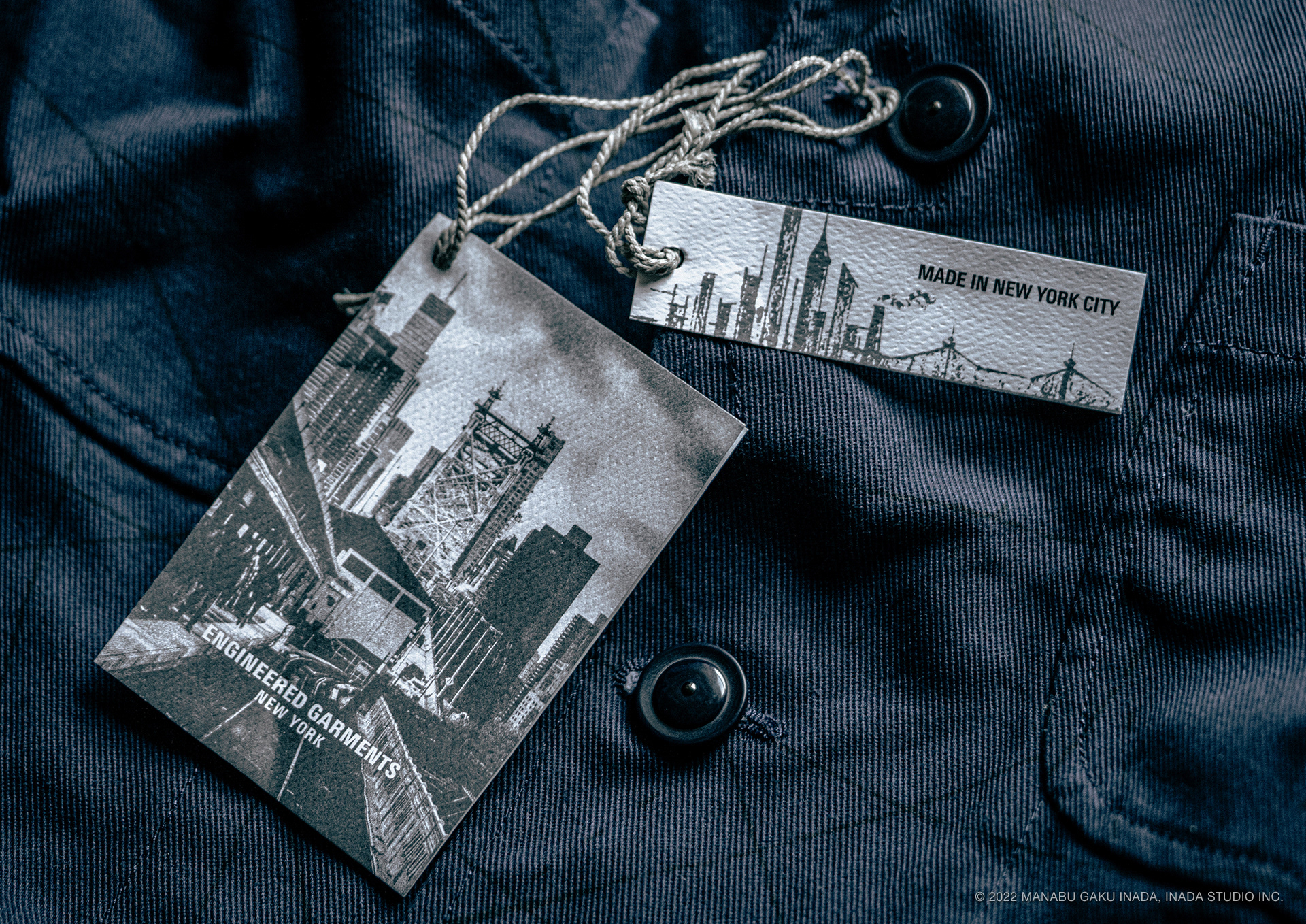 ENGINEERED GARMENTS Brand ID Brand Identity Development and Photography, 2022 by Manabu Gaku Inada. INADA STUDIO INC.  In the spring of 2021, Daiki Suzuki of Engineered Garments approached GAKU with a request to redesign the brand's hang tags — introducing new photography alongside a separate "Made in New York" tag. Drawing on the Nepenthes office in Long Island City as a conceptual anchor, the project took shape around a set of photographs: frontal, composed images of iconic structures such as the Queensboro Bridge and Queens Plaza.  Each image carried a private resonance. In the autumn of 1992, GAKU moved from Tokyo to New York to study. On the morning of his first day at an art school in Manhattan, a packed subway car rounded a bend — and Queens Plaza station suddenly opened up before him, the skyline gleaming in the distance. The feeling that surged through him then has never faded.  As an artist, GAKU had spent years photographing and sketching the city. For these tags, he drew from that personal archive, selecting each image with quiet intention. Together with a "Made in New York" tag — its design sparse and schematic, like a blueprint tracing the skyline — they were attached to every piece in the Spring/Summer 2022 collection.  Each tag rests in silence as part of the garment, holding a New York that belongs, in the end, to whoever is wearing it.  Brand:  Engineered Garments  Photography, Graphic Design:  Manabu Gaku Inada Year:  2022