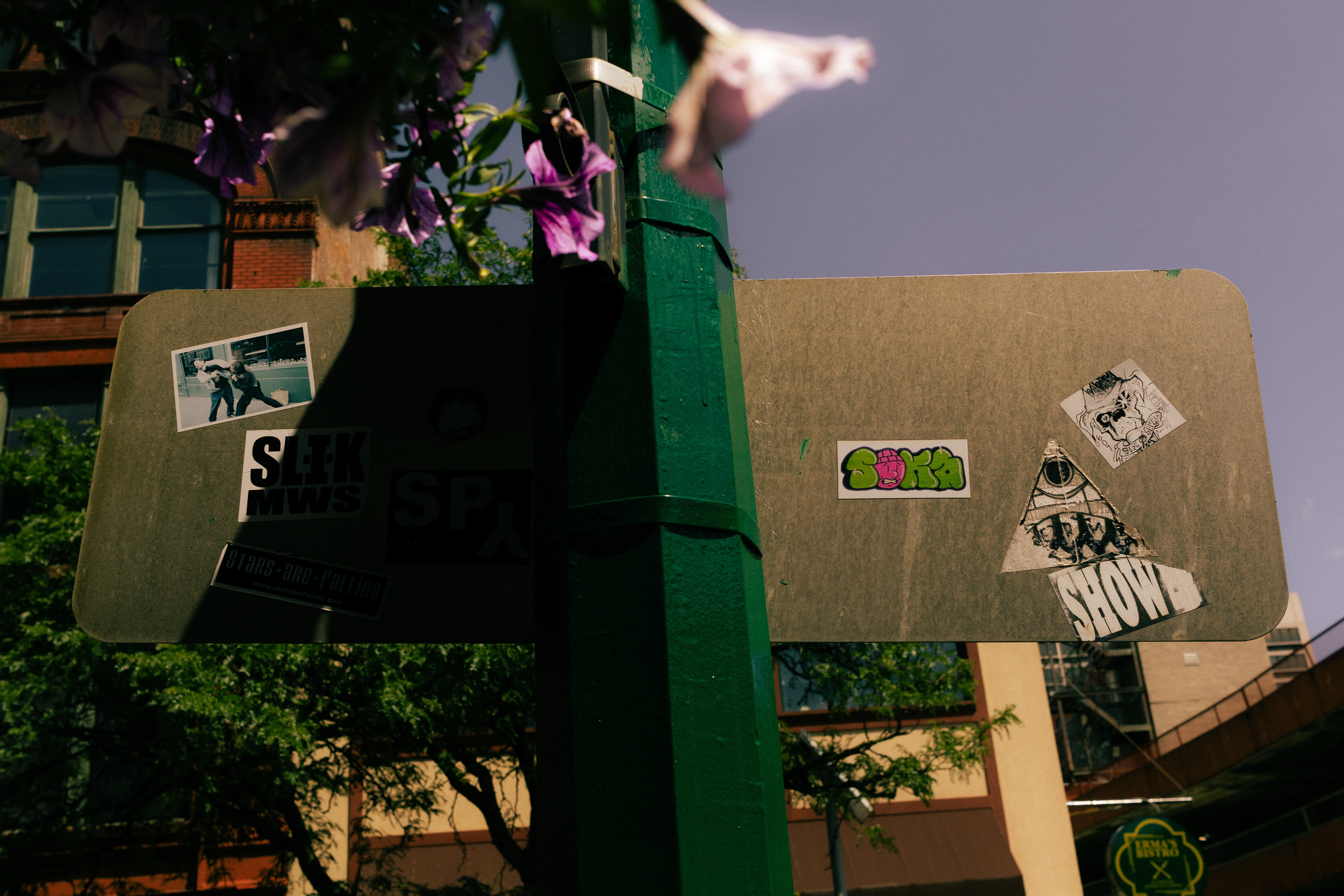 Sticker-covered street sign surrounded by greenery and colorful buildings.