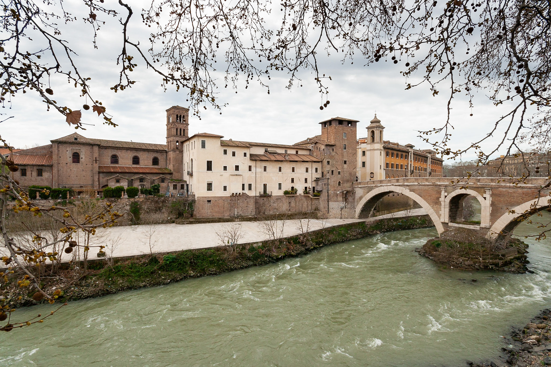 Isola Tiberina from the East Bank of the Tiber river framed by the bare branches of trees that line the pathway, Rome, Italy.