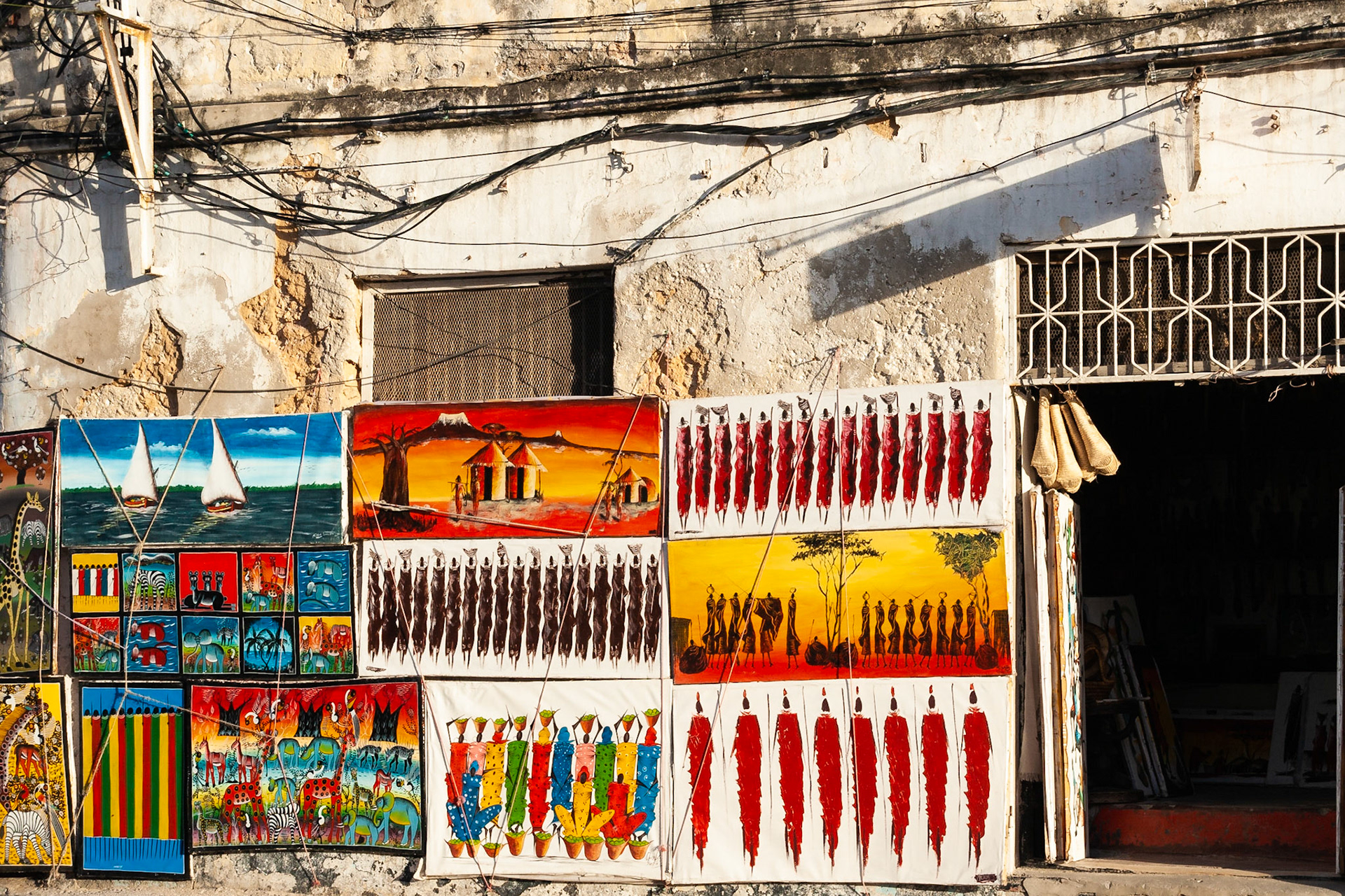 An outside wall serves as a gallery to display a selection of colourful paintings for sale, Stonetown, Zanzibar.