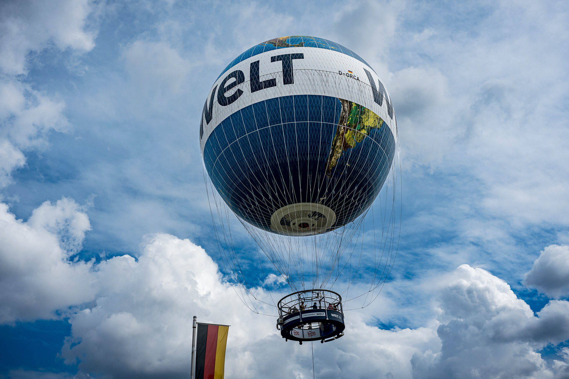 Low-angle shot of the Welt Balloon floating against a deep cobalt blue sky with white fluffy clouds. The balloon features a graphic map of the world. A small German flag is visible in the bottom corner.