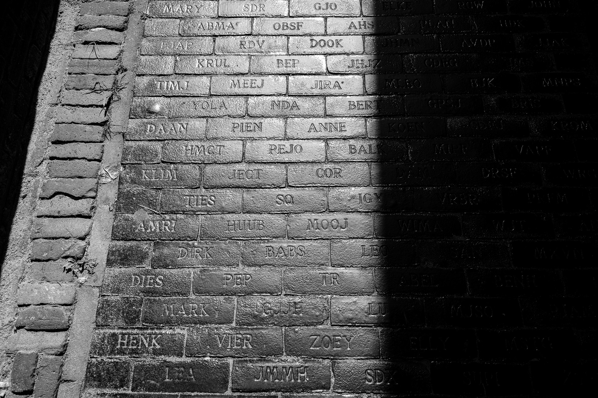 A high-contrast black and white architectural study of engraved names on cobblestones, showing deep inky shadows and tactile stone textures achieved with a Sony A7CR and Leica Monochrom CCD emulation.