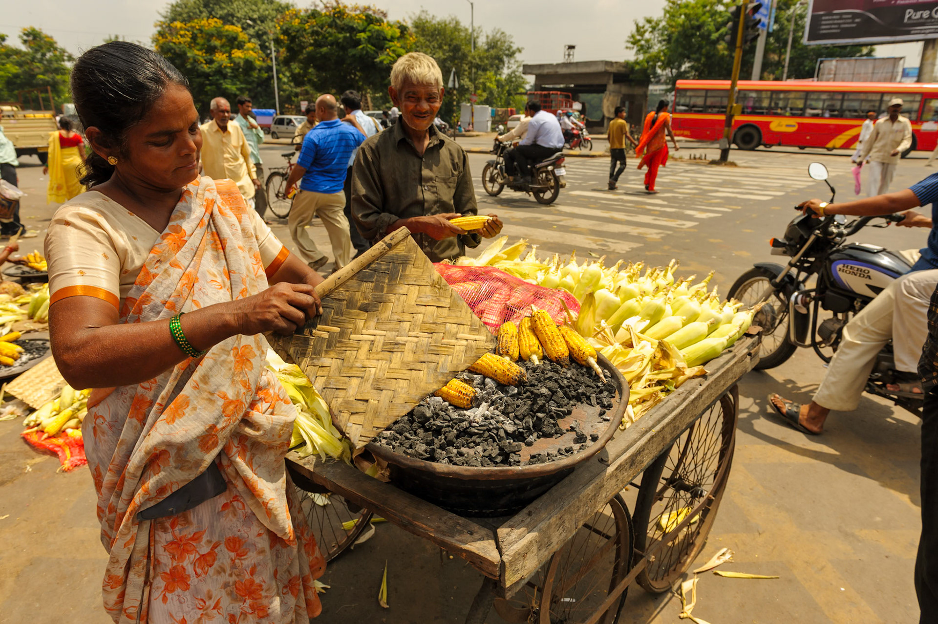 Pune, India 9/18/2010 --- An Indian street corn seller turns the corn from the fire while fanning using a traditional fan.  Her elderly father is helping on the side.  Grilled sweet corn on the cob is one of the typical street food in India.