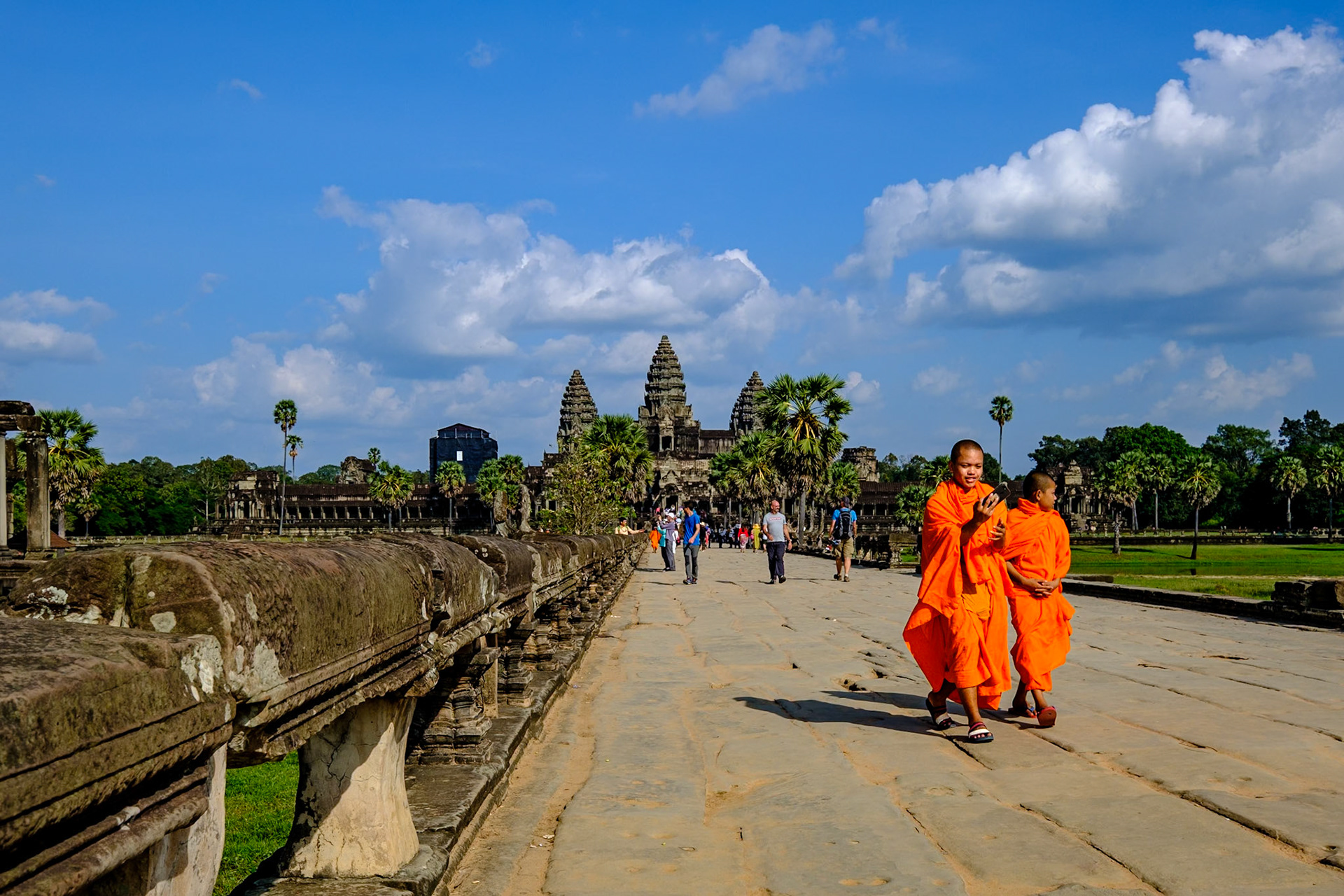 This vibrant photograph showcases the iconic Angkor Wat temple complex in Siem Reap, Cambodia, a UNESCO World Heritage site. In the foreground, two Buddhist monks in bright orange robes walk along the ancient stone pathway, providing a lively contrast to the historical setting. The temple's distinctive towers rise majestically in the background, framed by blue skies and lush greenery. Tourists explore the site, underscoring Angkor Wat's significance as a major global attraction. The image captures the serene and timeless beauty of this architectural marvel, blending history, culture, and spirituality.