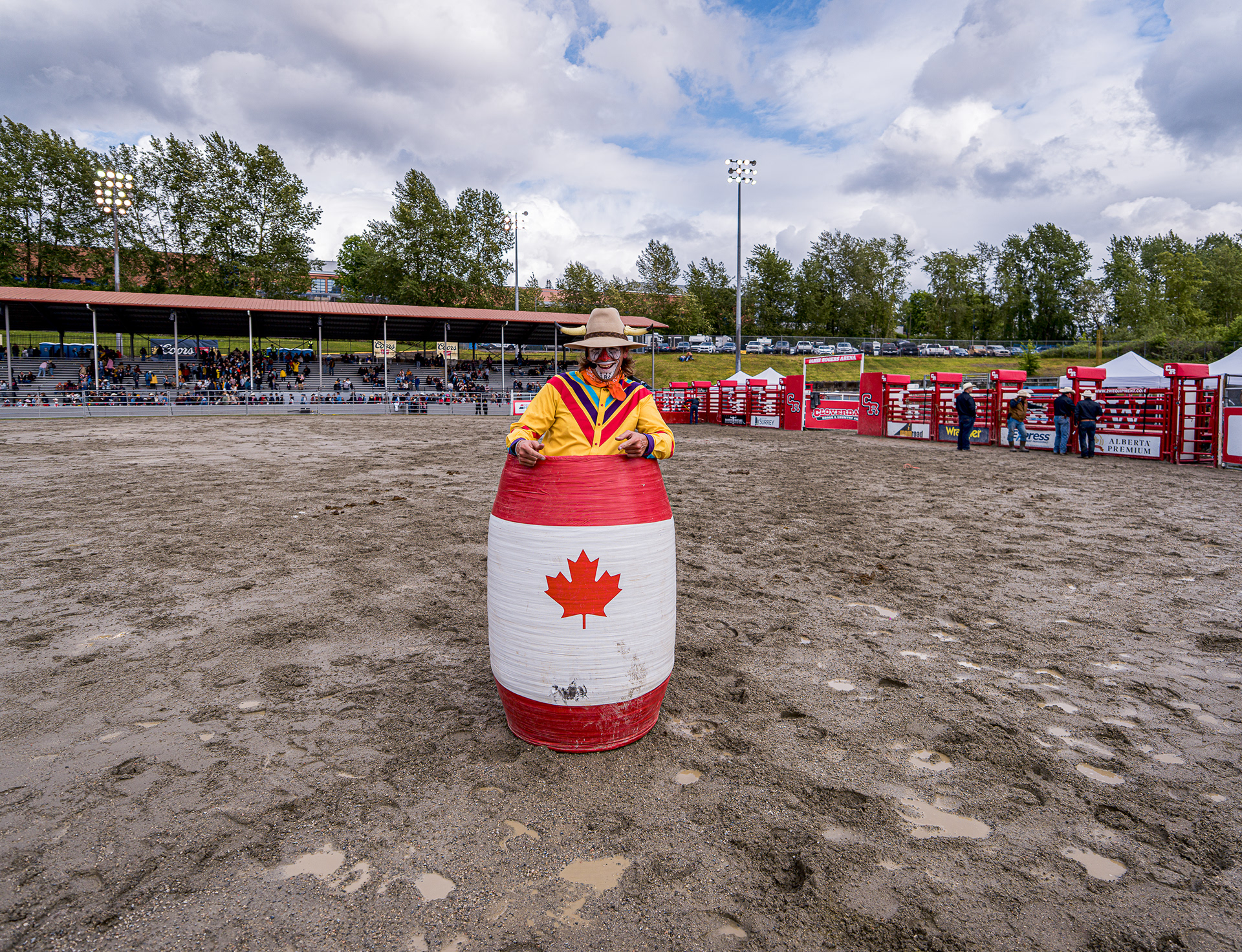 A rodeo clown standing inside a barrel painted with the Canadian flag in the middle of a dirt arena.