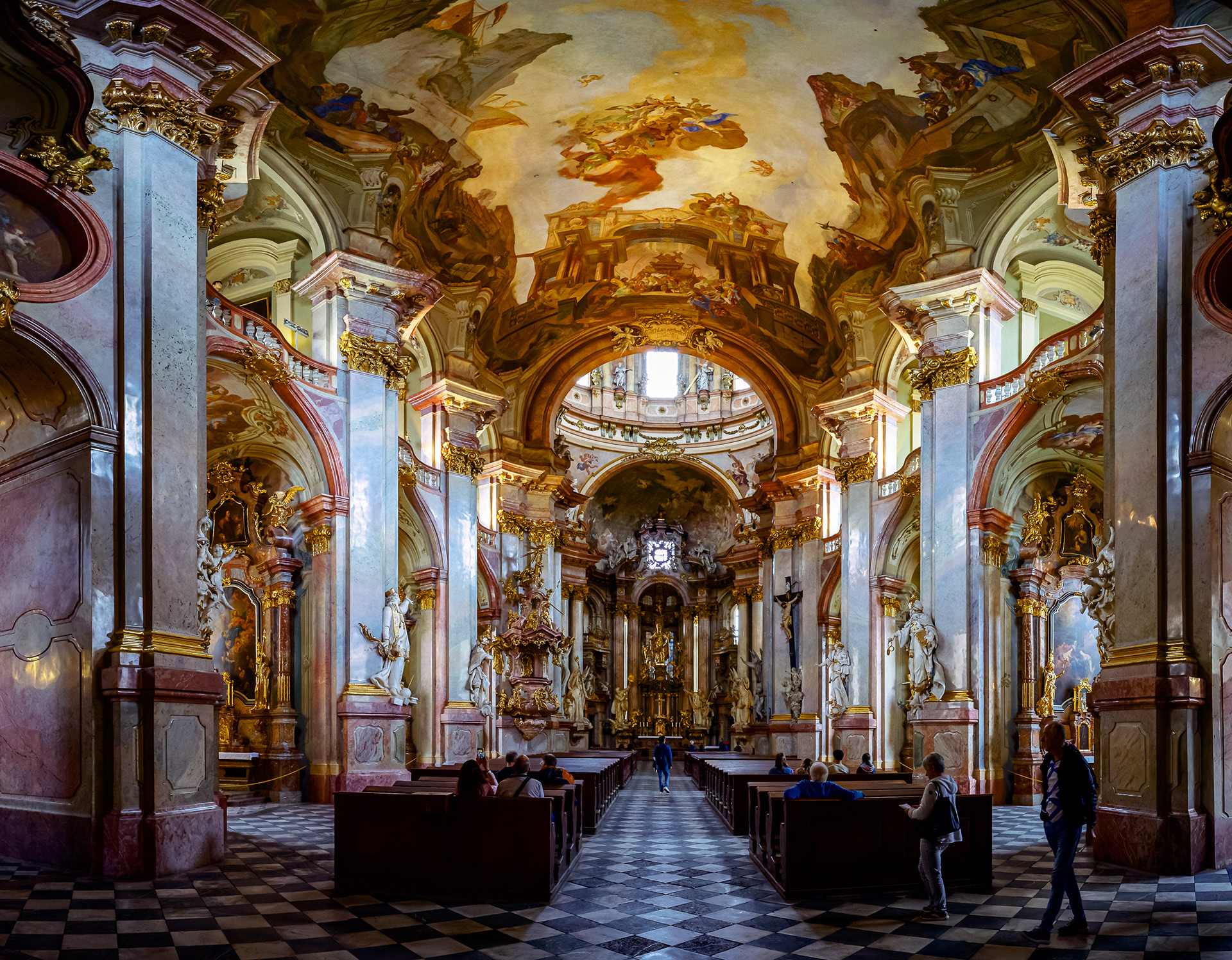Vertical panorama of the St. Nicholas Church interior in Prague, showing massive marble columns and a gold ceiling fresco.