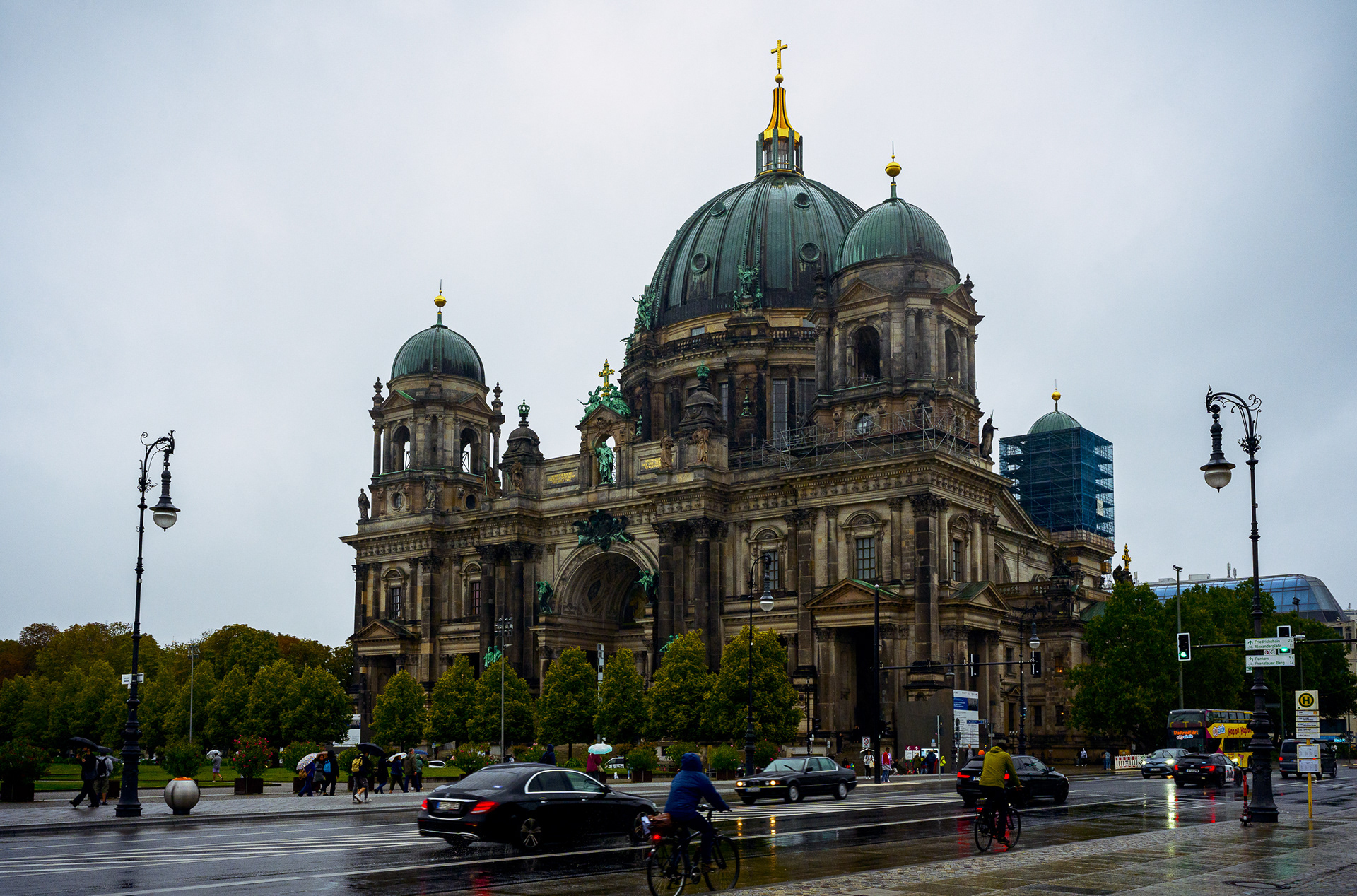 Cinematic shot of the Berlin Cathedral (Berliner Dom) on a rainy day. The wet asphalt of the street acts as a mirror, reflecting the green dome and the trees. Pedestrians with umbrellas walk through the moody, atmospheric scene.