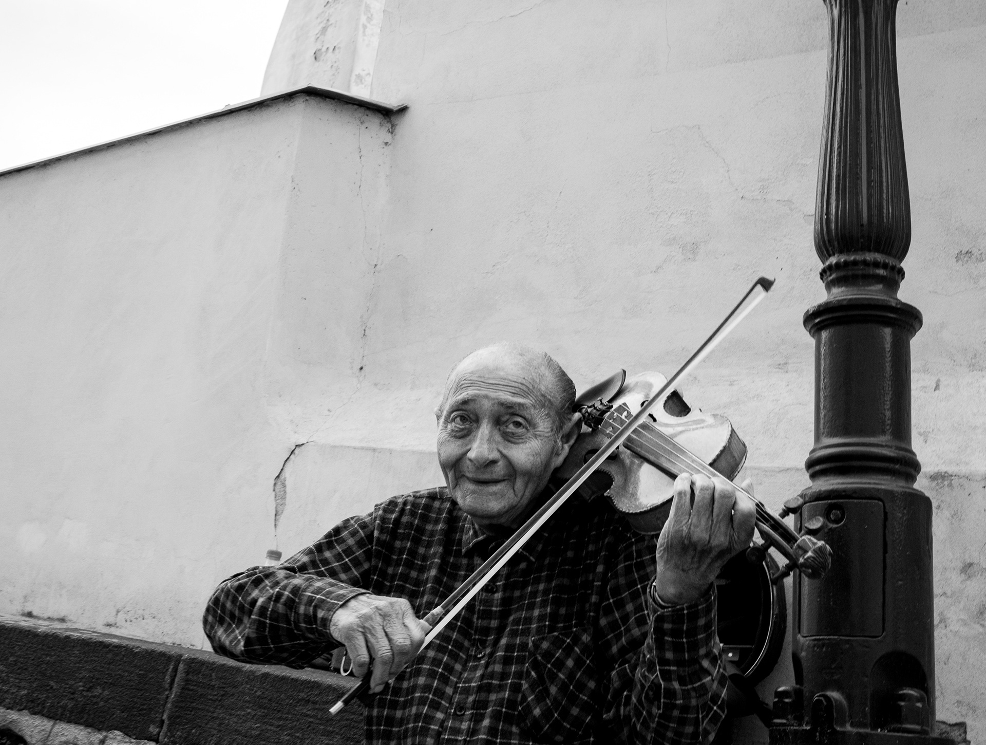 A 4:5 black and white street portrait of an elderly violinist, captured on a Sony A1 with Leica Monochrom CCD Yellow Filter emulation for radiant skin tones and high-key spectral luminance.