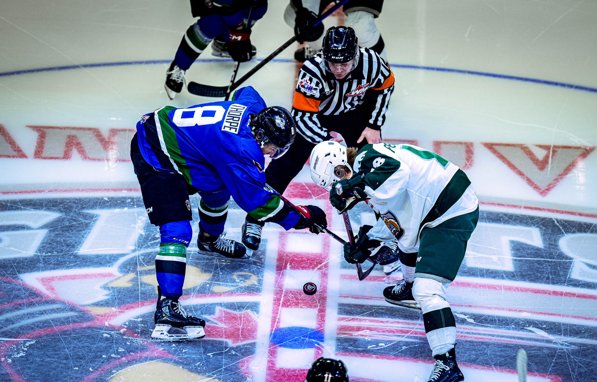 A dramatic high-angle overhead sports photograph capturing the precise moment of a WHL face-off between the Vancouver Giants and Everett Silvertips at the Langley Events Centre in Langley, BC. The composition highlights the fierce geometry of crossed sticks over the puck and the intricate texture of the scratched ice surface. Captured on the Sony A1 by Joe Ng, utilizing Leica M9 color science for weighted tonal depth.