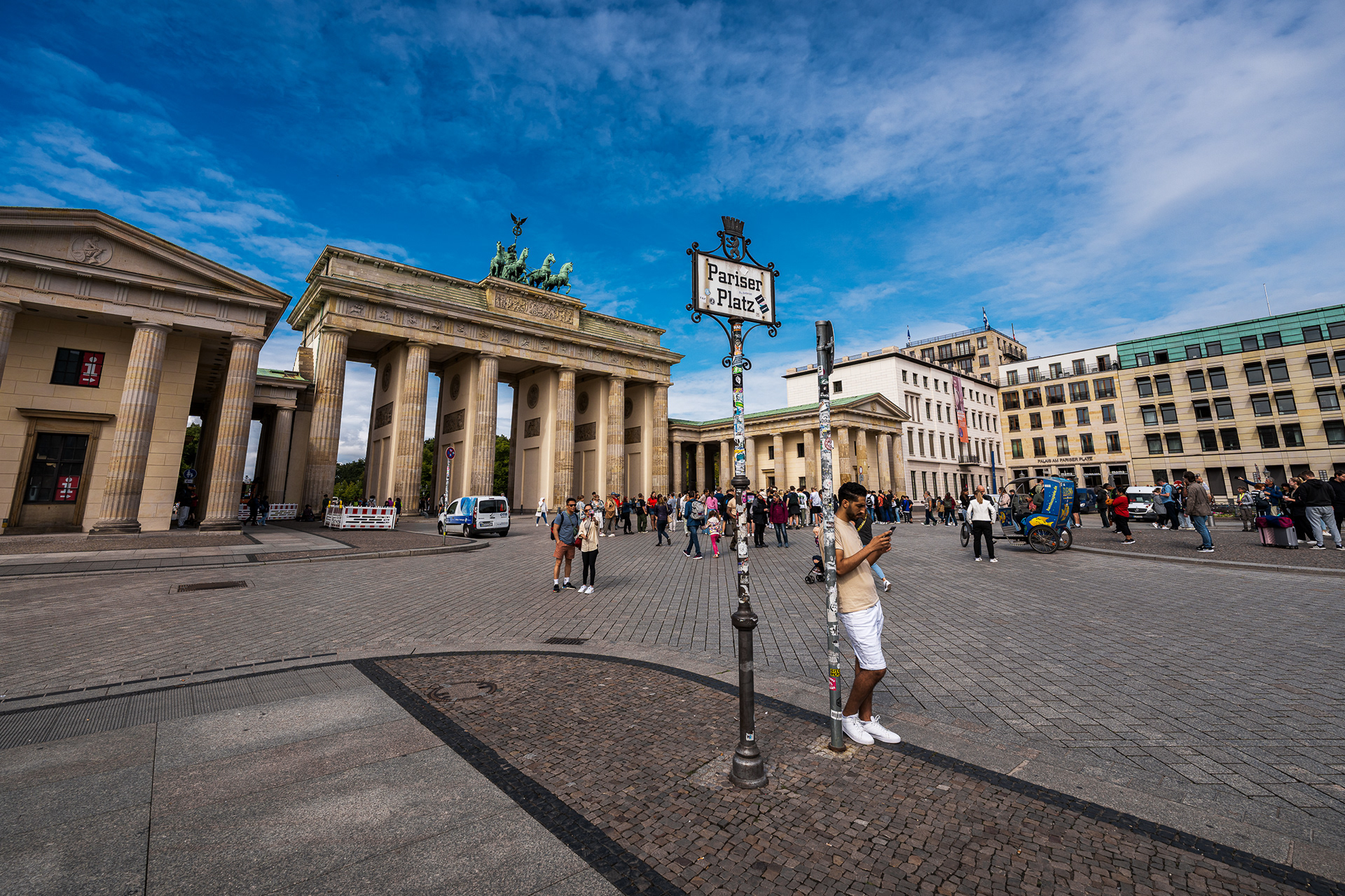 Wide-angle architectural photograph of the Brandenburg Gate in Berlin under a deep blue summer sky. Tourists walk on the cobblestones of Pariser Platz, dwarfed by the massive sandstone columns and the Quadriga statue on top.