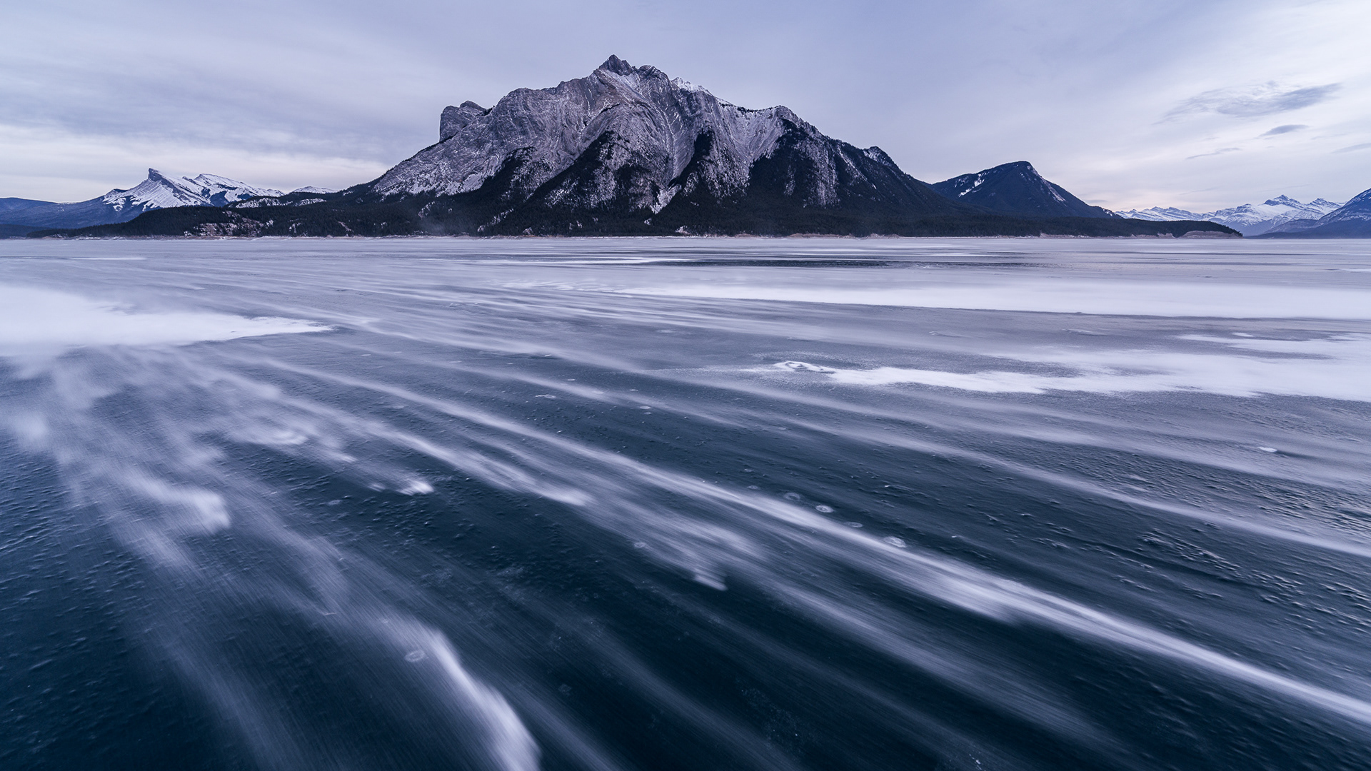 Cinematic long-exposure landscape of wind-swept snow across the frozen surface of Abraham Lake, Alberta. Dramatic mountain peaks under a moody winter sky, shot on Sony A7CR 61MP by Joe Ng.