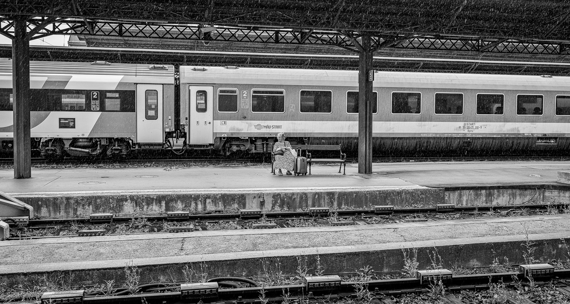A solitary woman sitting on a bench on a rain-slicked train platform in Budapest.