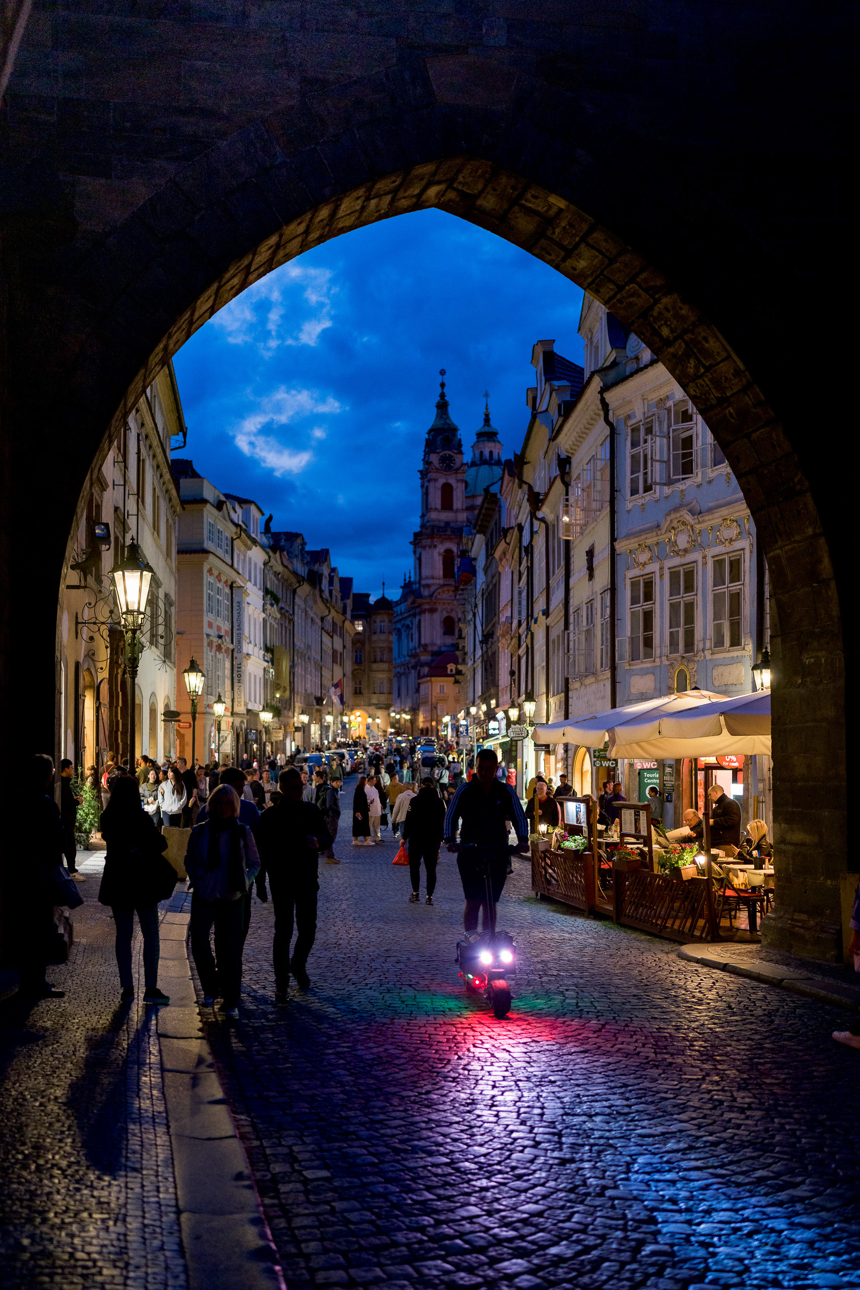 Blue hour street scene in Prague framed by a gothic stone archway, contrasting warm street lights with a deep blue sky.