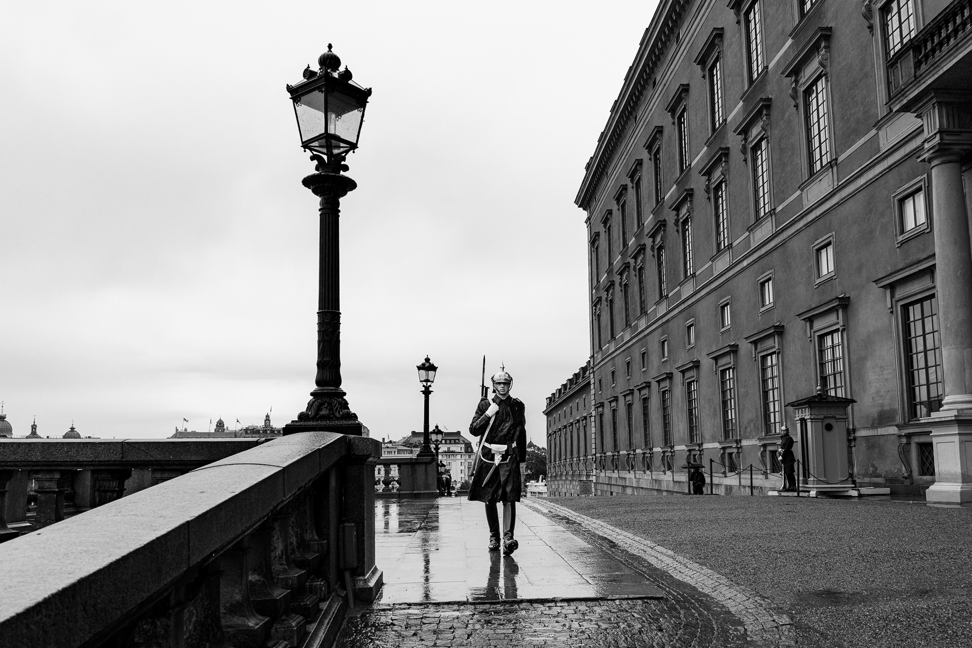 A high-contrast black and white street photo of a royal guard walking past the Stockholm Palace, processed with Leica M10 Monochrom emulation on a Sony A1 to emphasize specular highlights on wet pavement.